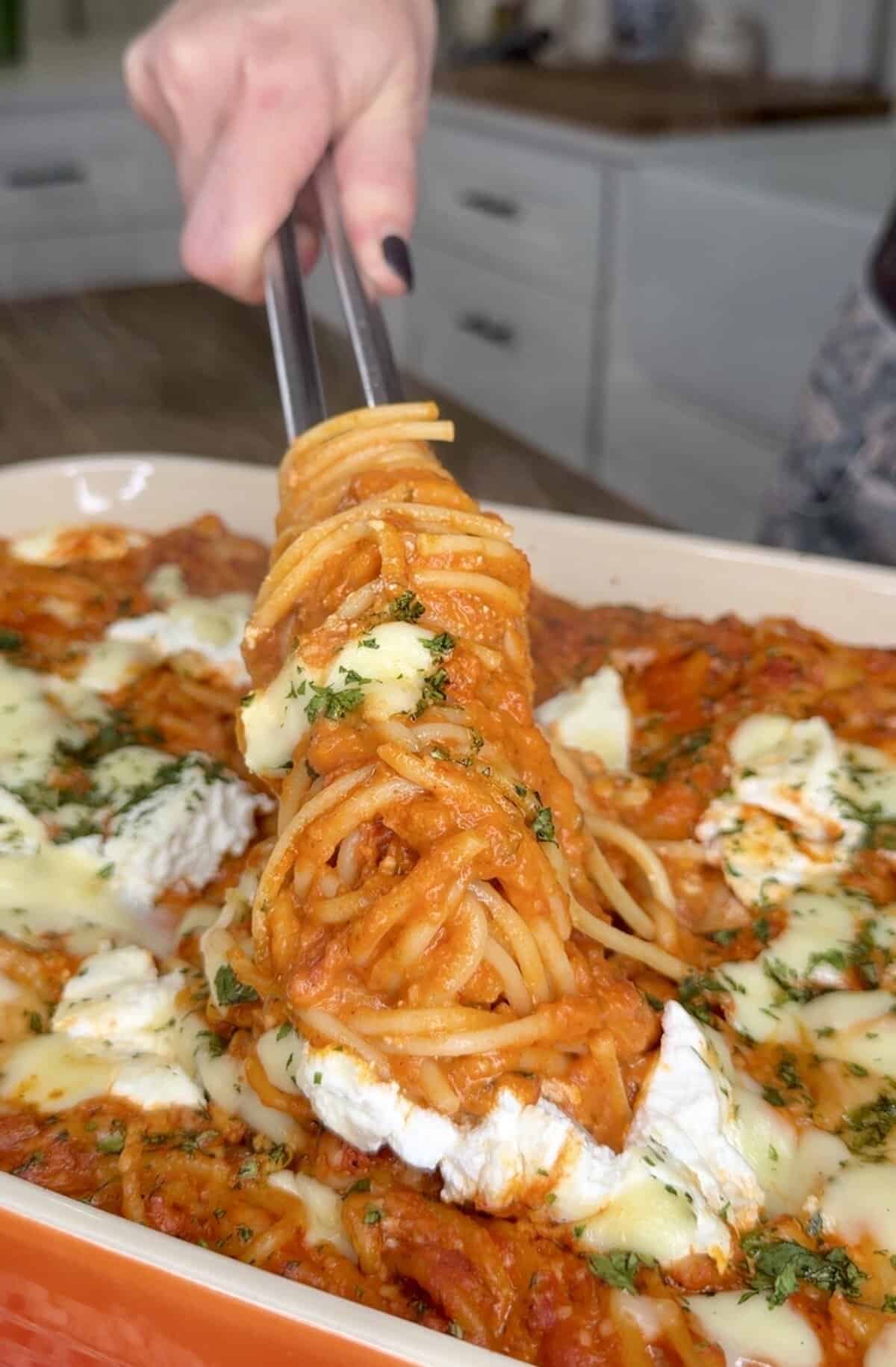 A hand uses tongs to lift a serving of cheesy baked spaghetti with tomato sauce and herbs from a casserole dish in a kitchen setting.