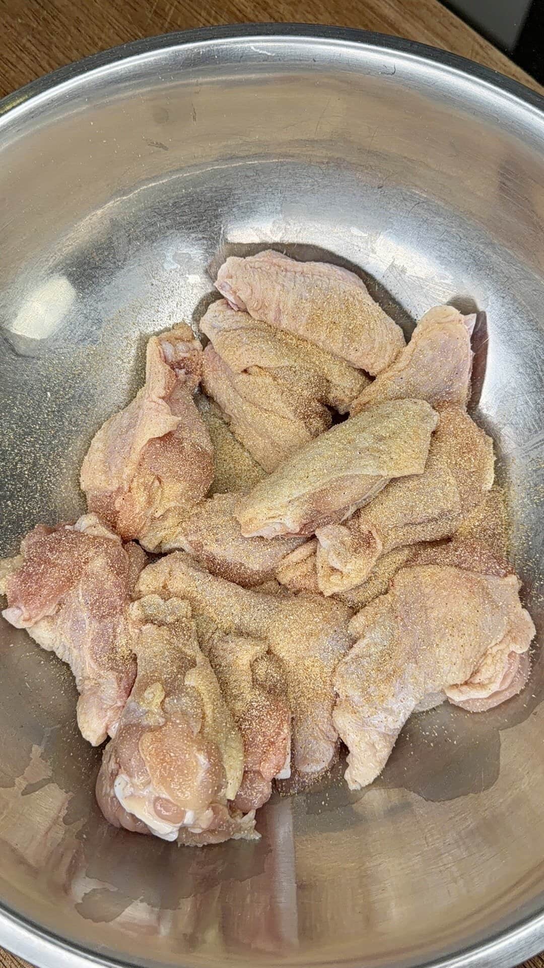 Raw chicken wings in a large metal bowl, sprinkled with a generous amount of dry seasoning powder, ready to be mixed or cooked into delicious crispy oven-baked wings.