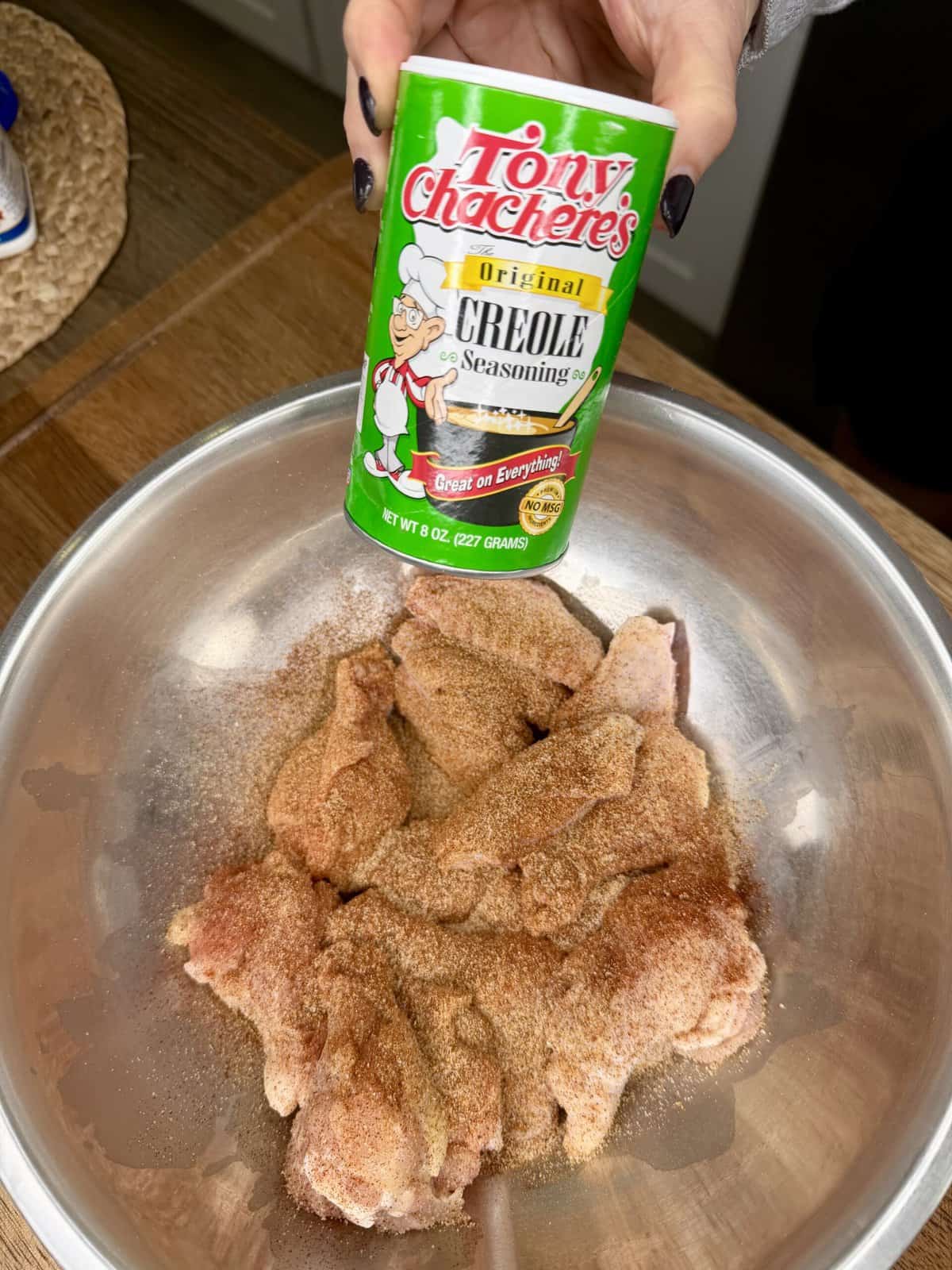 A hand holds a can of Tony Chachere’s Creole Seasoning above a metal bowl filled with raw chicken drumsticks, perfectly seasoned and ready to be transformed into crispy oven-baked wings.
