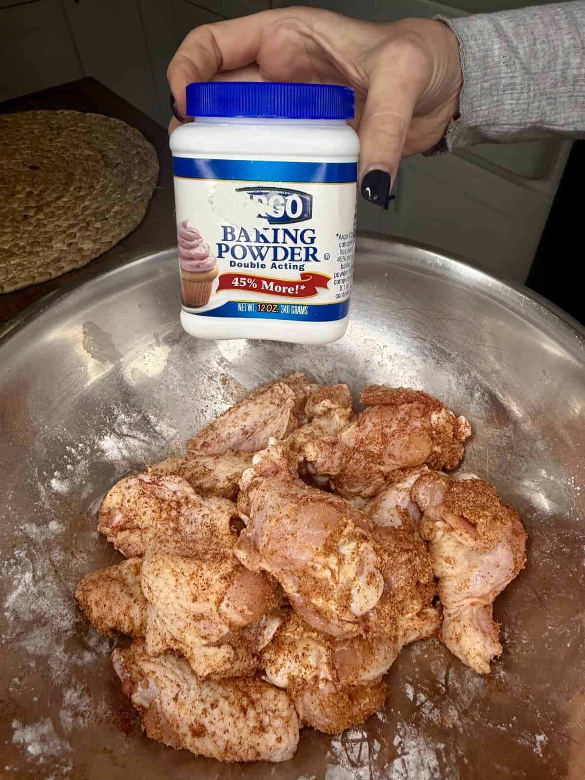 A hand holds a container of Argo baking powder above a metal bowl filled with raw, seasoned chicken wings—perfect for making crispy oven-baked wings in a kitchen setting.