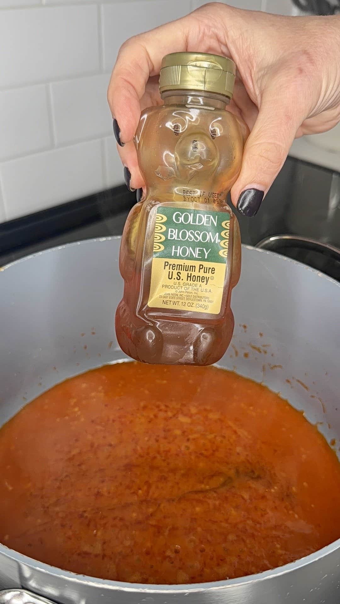 A hand holds a bottle of Golden Blossom honey above a pot of simmering tomato sauce on a stovetop, perfect for drizzling over Crispy Oven-Baked Wings. White tile backsplash is visible in the background.