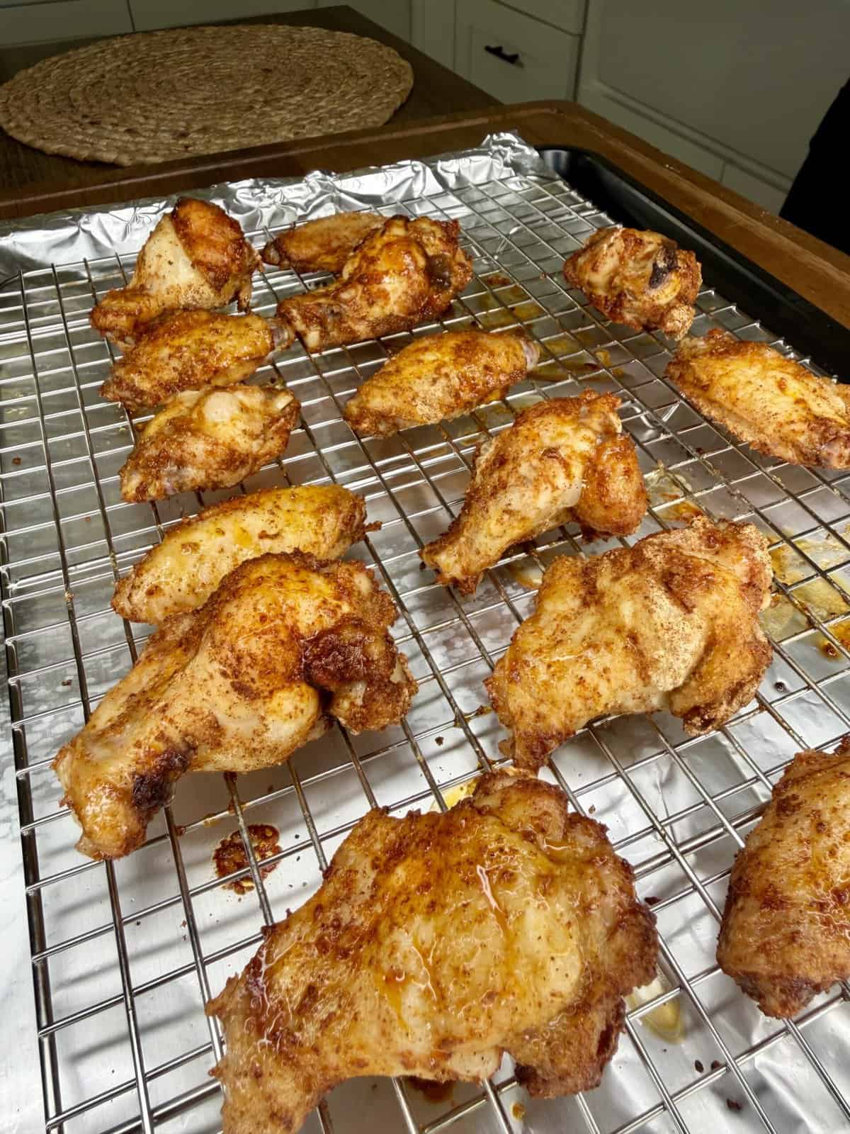 Crispy Oven-Baked Wings coated in seasoning are arranged on a metal wire rack over a foil-lined baking sheet, ready for or fresh out of the oven. A kitchen countertop and cabinets can be seen in the background.