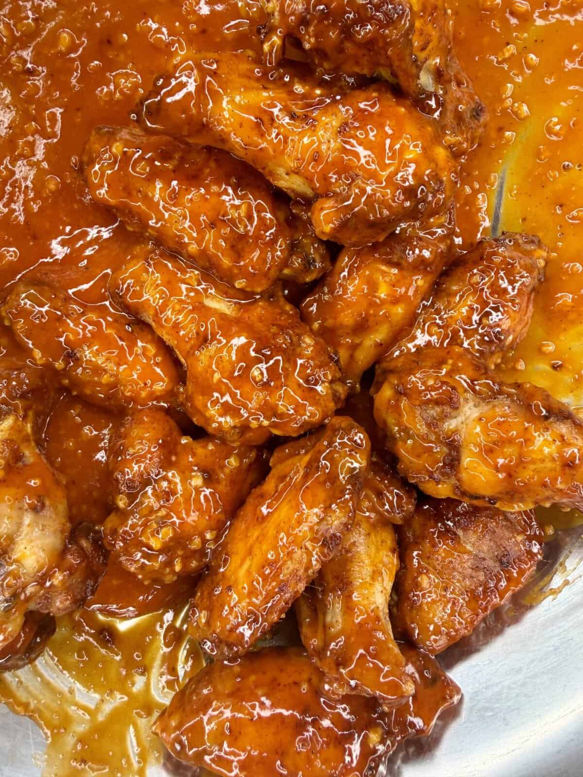 Close-up of several crispy oven-baked wings coated in a thick, glossy, orange-red hot sauce, arranged in a metal tray with sauce pooling around them.