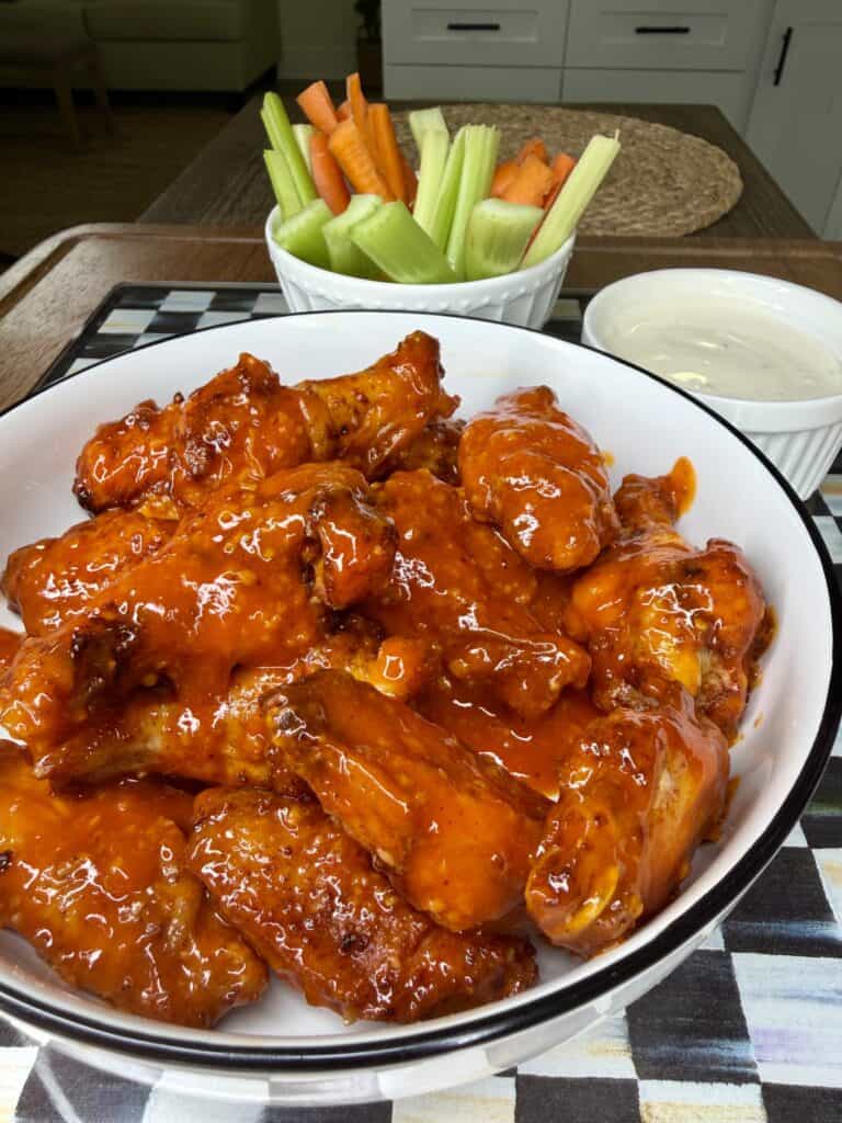 A bowl of saucy Crispy Oven-Baked Wings sits on a table alongside a cup of ranch dressing and a bowl of celery and carrot sticks for dipping.