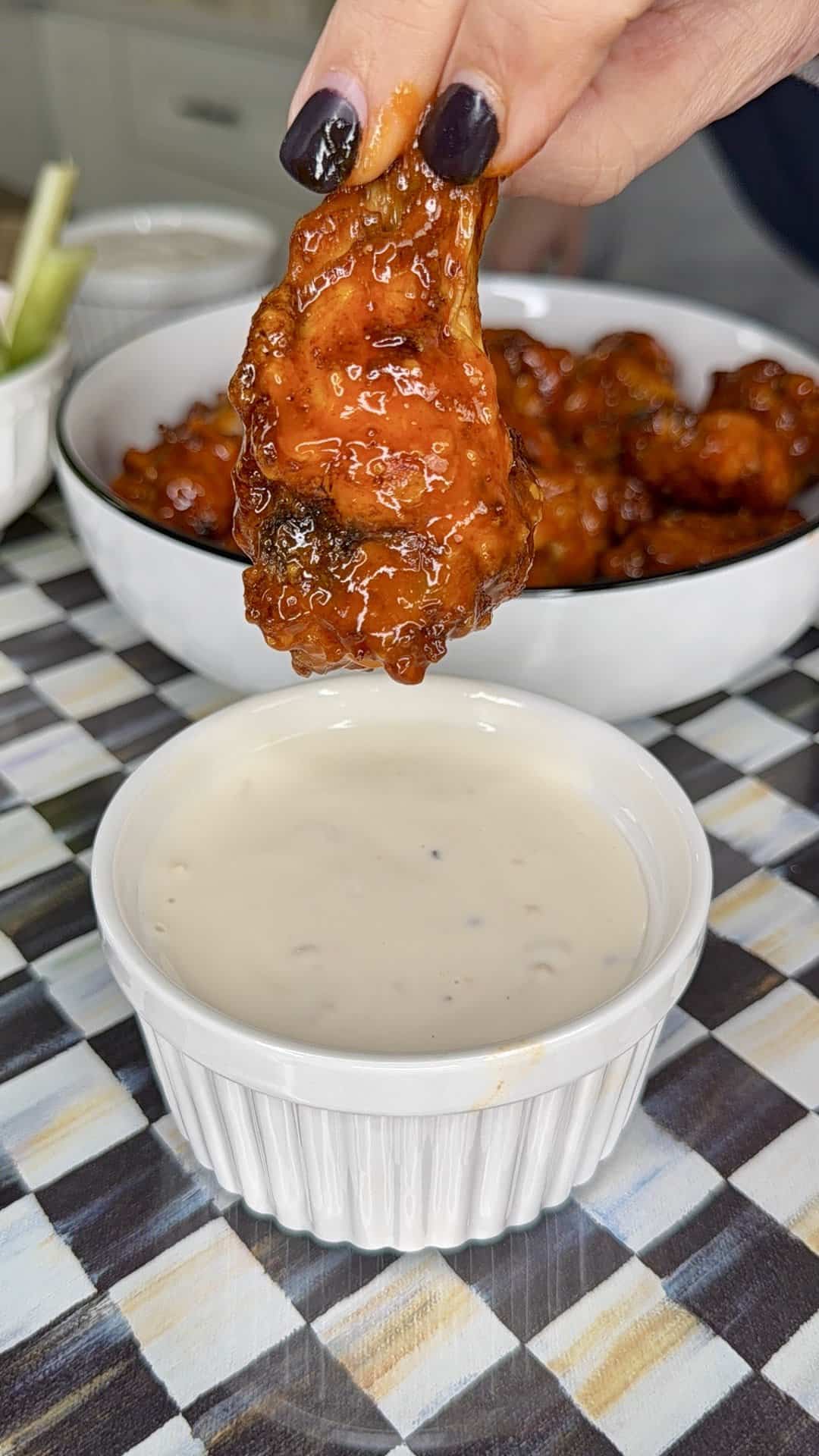 A hand with dark nail polish holds a saucy Crispy Oven-Baked Wing above a ramekin of creamy dipping sauce, with a bowl of more chicken wings in the background on a checkered tablecloth.