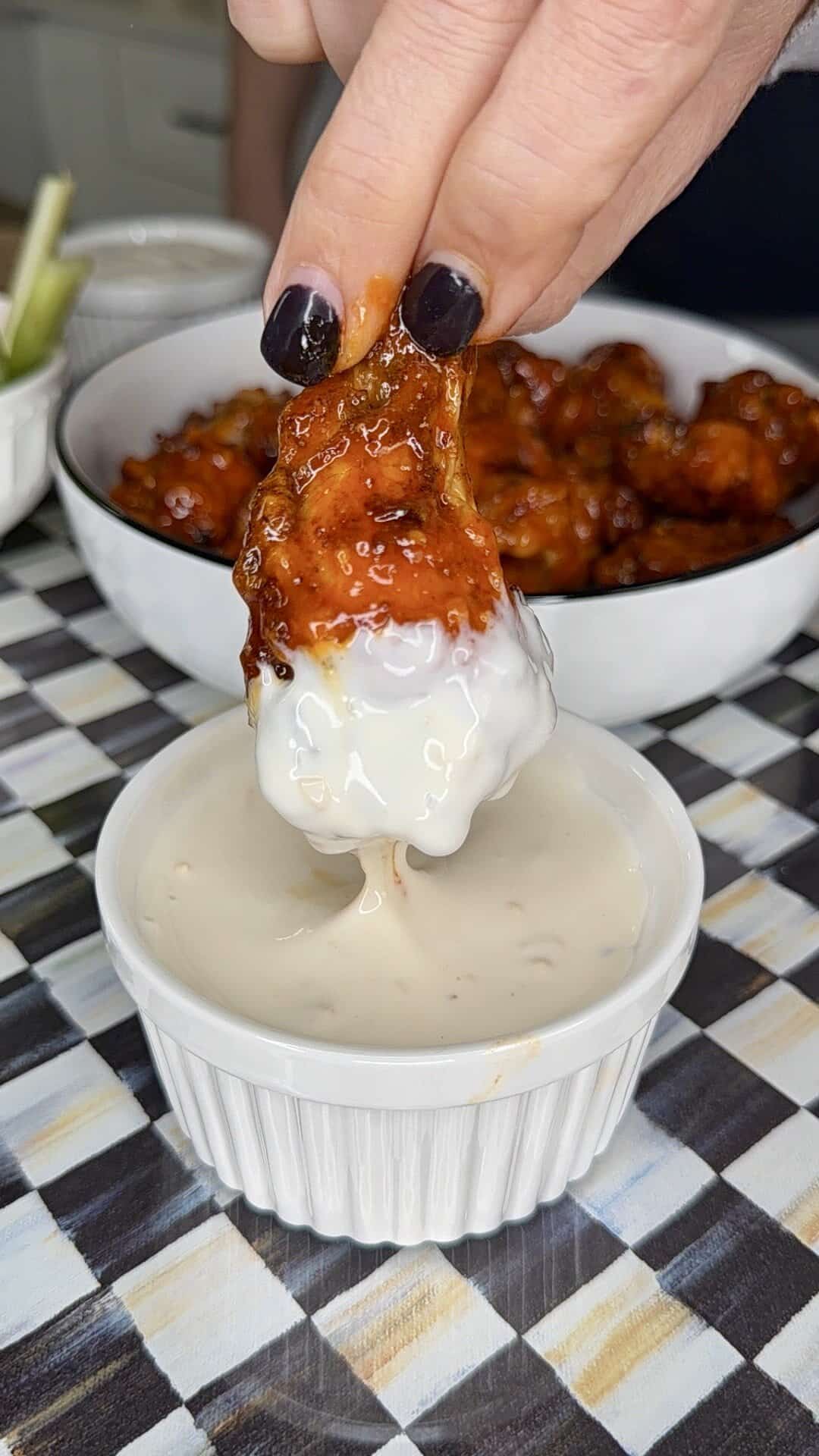 A hand with dark nail polish dips a saucy Crispy Oven-Baked Wing into creamy ranch dressing, with more wings and celery sticks in the background on a checkered tablecloth.