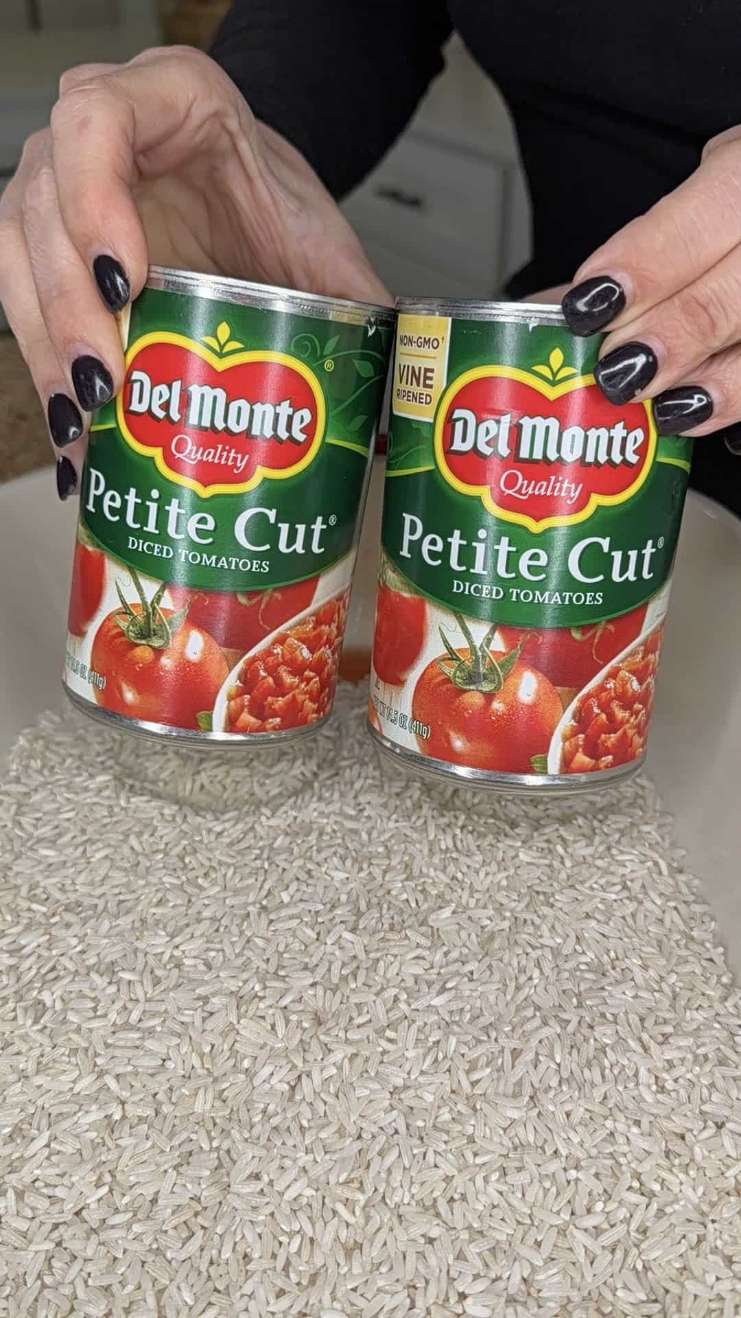 Two hands with black-painted nails hold cans of Del Monte Petite Cut diced tomatoes over a bowl filled with uncooked white rice.