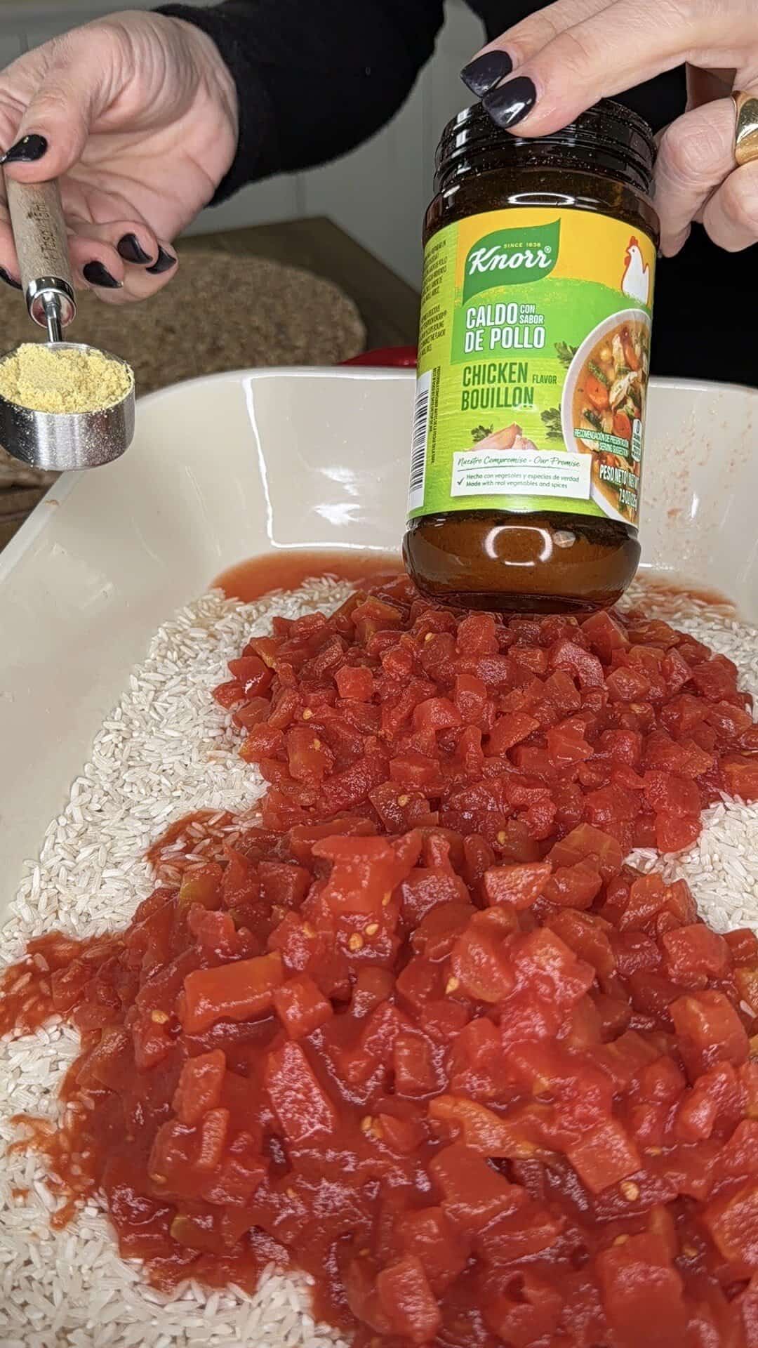 A person holds a jar of Knorr chicken bouillon and pours a spoonful into a dish filled with uncooked rice and diced tomatoes.