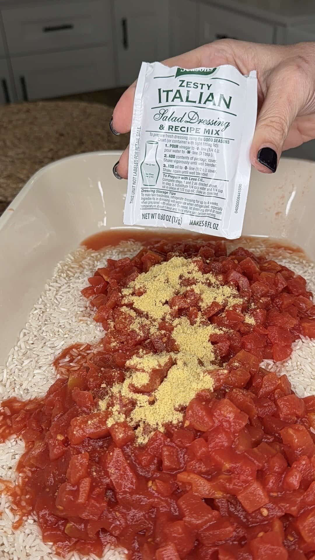 A hand holds a packet of Zesty Italian salad dressing mix above a baking dish filled with uncooked rice, diced tomatoes, and seasonings, ready to be mixed.