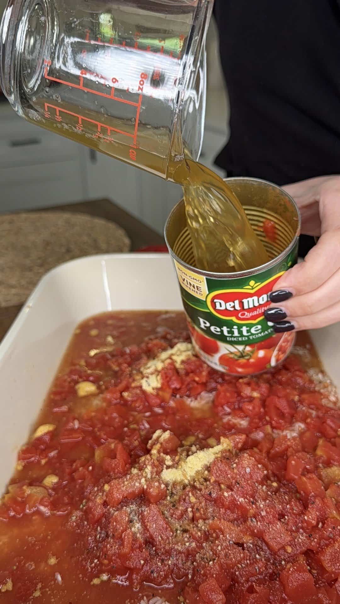 A person pours liquid from a measuring cup into an empty Del Monte Petite Diced Tomatoes can over a dish filled with canned tomatoes and seasonings, preparing a recipe.