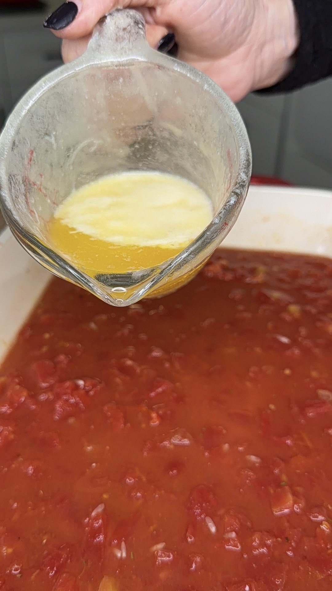 A hand pours melted butter from a glass measuring cup into a dish filled with chunky tomato sauce.
