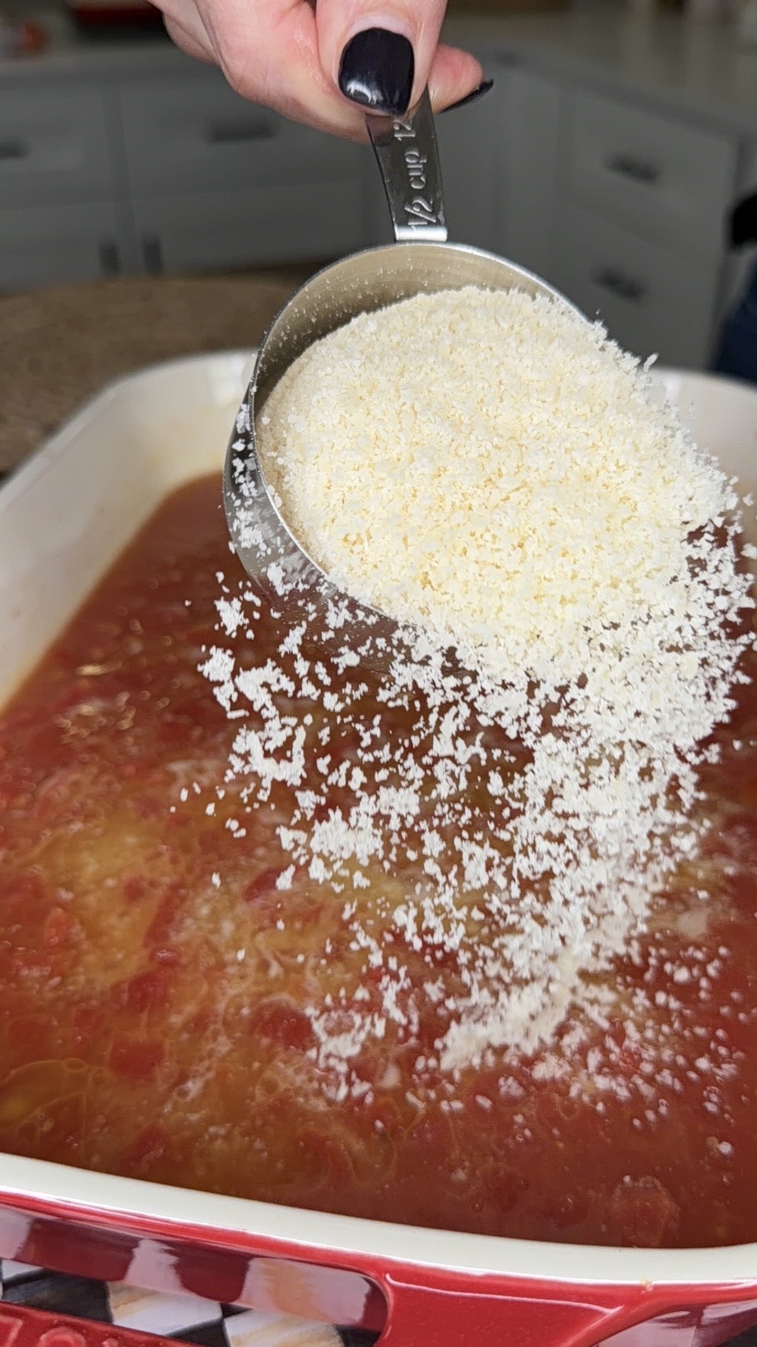 A hand holds a metal measuring cup, pouring grated Parmesan cheese into a casserole dish filled with a tomato-based mixture. Kitchen counters and cabinets are blurred in the background.