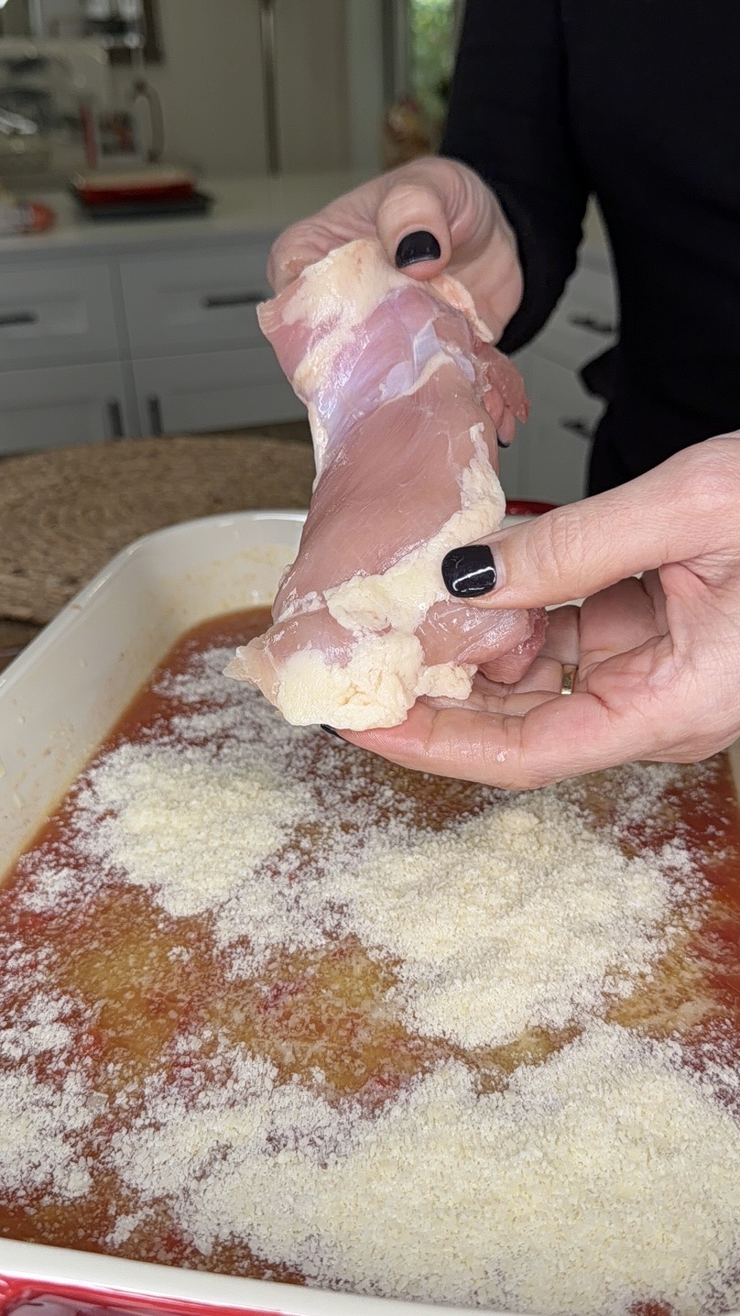 A person with black nail polish holds a raw chicken thigh over a casserole dish filled with tomato sauce and sprinkled with grated cheese, preparing to add the chicken to the dish.