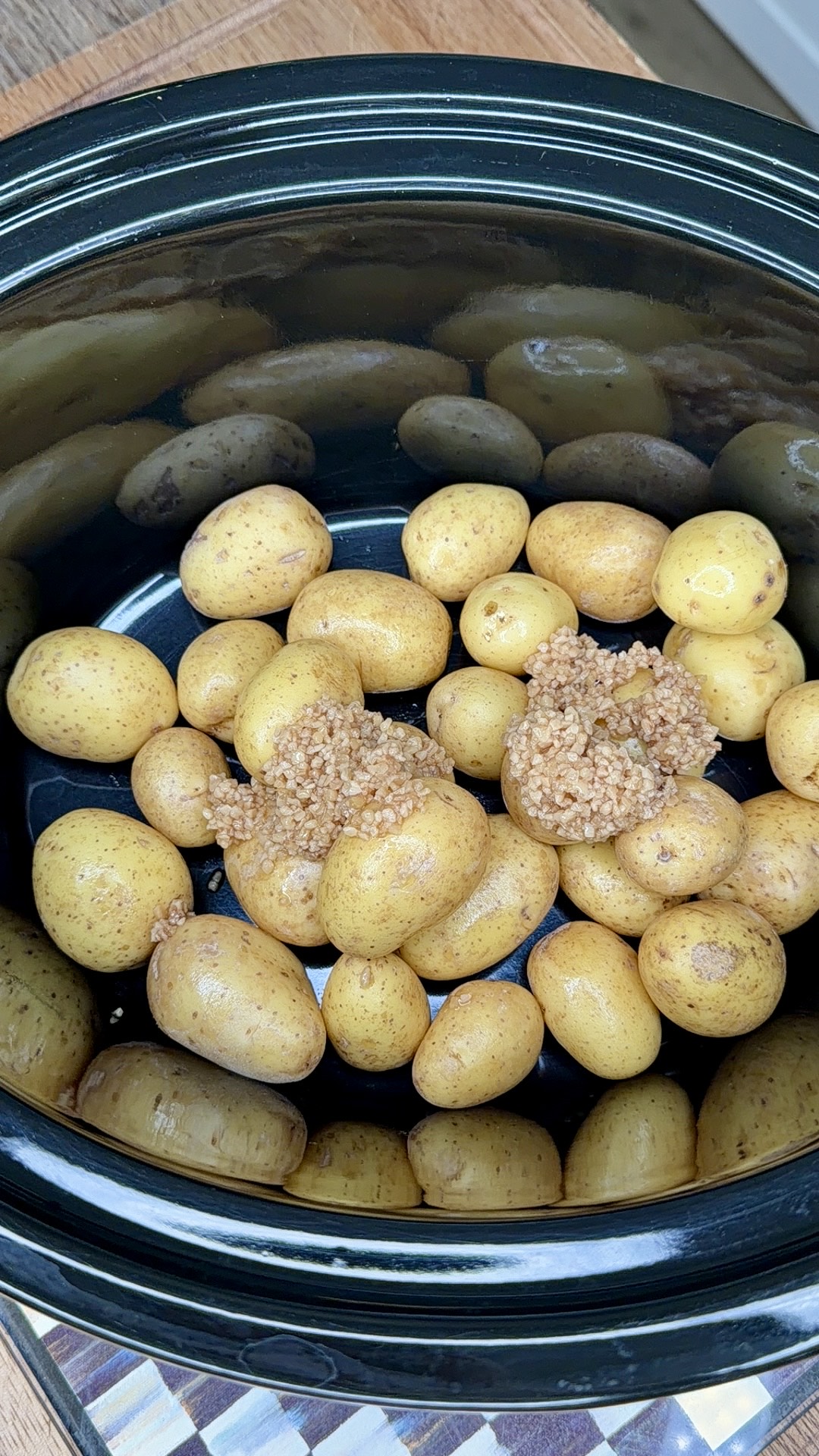 Small yellow potatoes with two spoonfuls of minced garlic sit at the bottom of a black slow cooker, ready to be cooked.