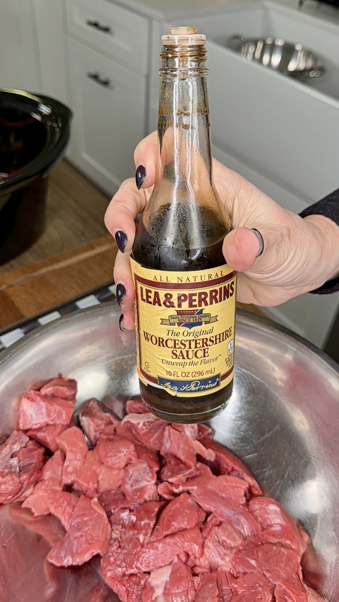 A hand with dark nail polish holds a bottle of Lea & Perrins Worcestershire Sauce over a metal bowl filled with raw beef slices in a kitchen setting.