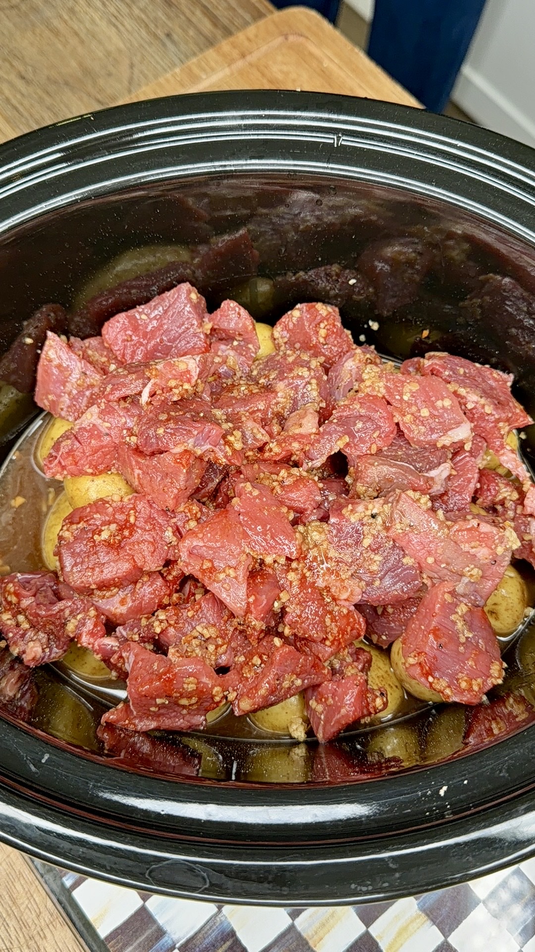 Chunks of raw seasoned beef are placed on top of halved baby potatoes inside a black slow cooker, ready to be cooked. The ingredients rest on a wooden countertop.