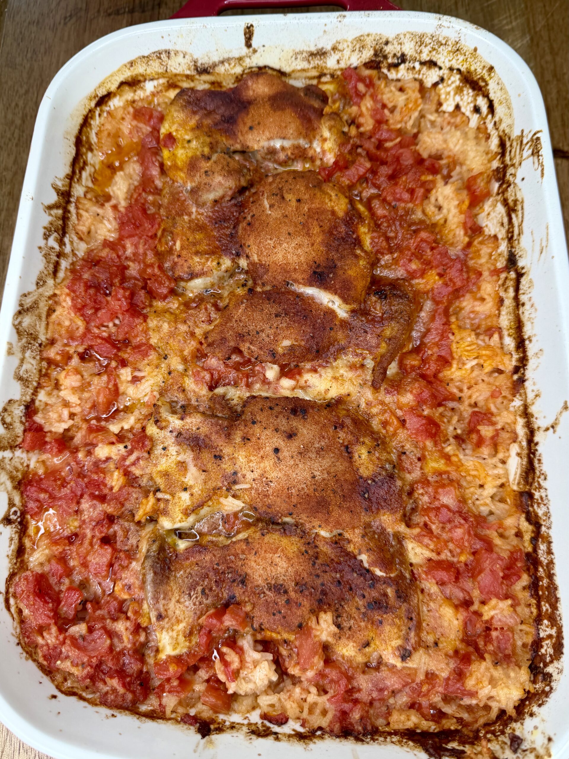 A white baking dish filled with baked chicken thighs, seasoned and browned, sitting on a bed of tomato and rice mixture. The edges of the dish show some browning from cooking.