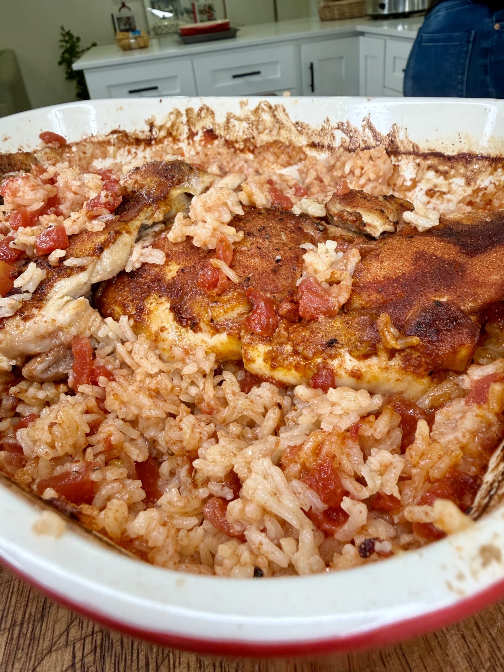 A close-up of a casserole dish filled with baked rice, tomatoes, and seasoned chicken. The top is golden brown, and some rice appears fluffed and moist with pieces of tomato mixed in. The dish sits on a wooden surface.