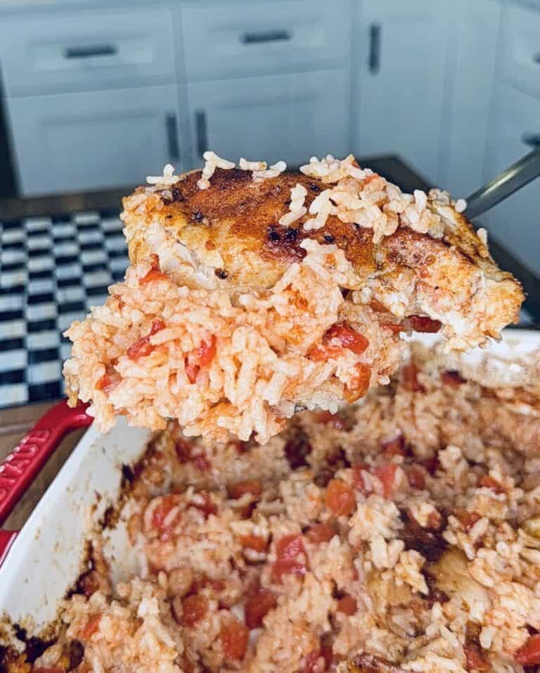 A close-up of a serving spoon lifting a portion of baked rice and chicken with tomatoes from a casserole dish in a bright kitchen.