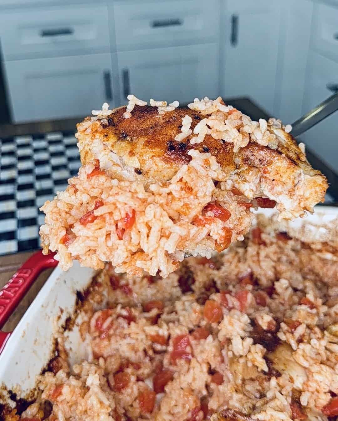 A close-up of a serving spoon lifting a portion of baked rice and chicken with tomatoes from a casserole dish in a bright kitchen.