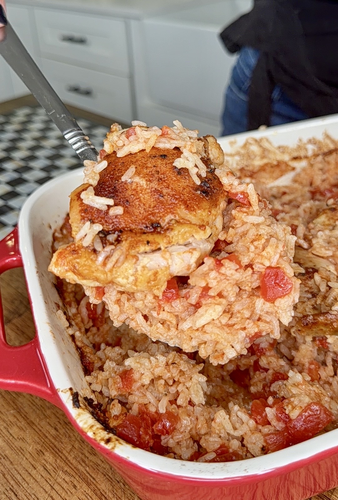 A close-up of a spoon lifting a serving of baked chicken with rice and tomatoes from a red casserole dish in a kitchen.