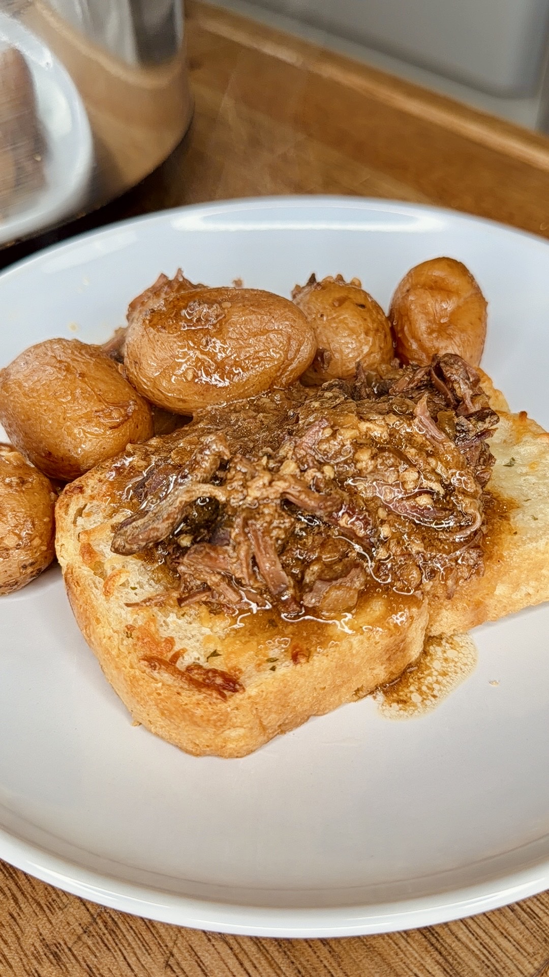 A white plate with roasted baby potatoes and a slice of toasted bread topped with shredded beef and gravy, set on a wooden surface.