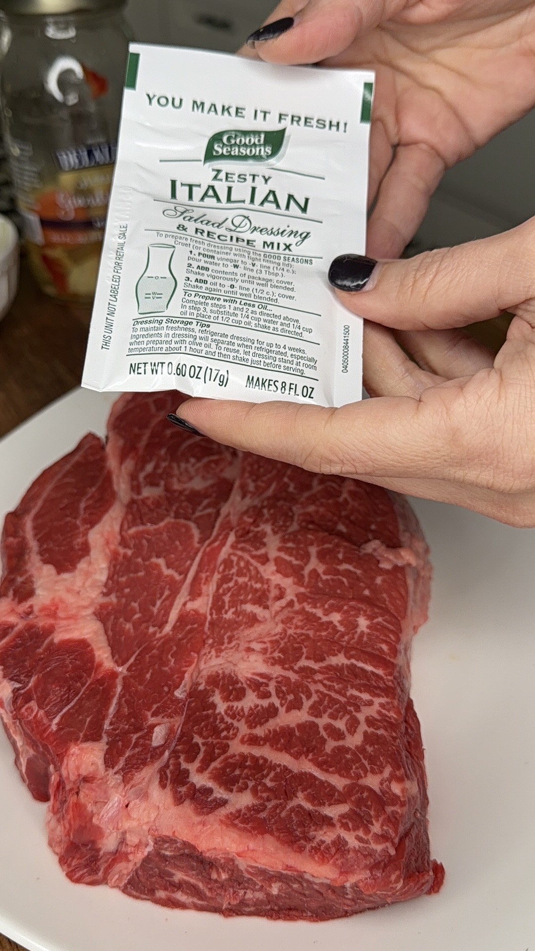 A hand holds a packet of Good Seasons Zesty Italian salad dressing mix above a raw, marbled steak resting on a white plate.