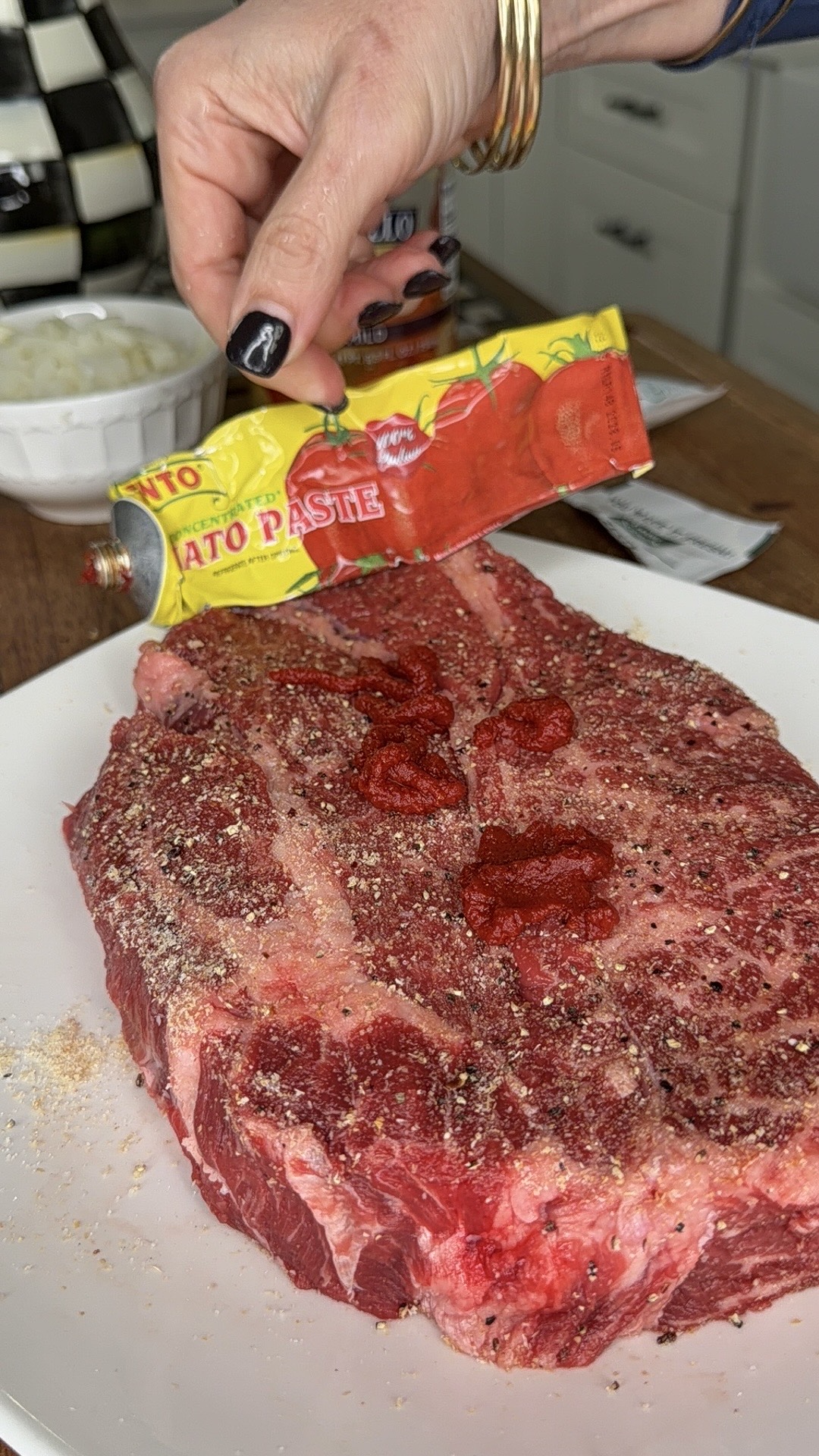 A hand squeezes tomato paste from a tube onto a seasoned raw steak on a white plate, with a checkered container and chopped onions in the background.