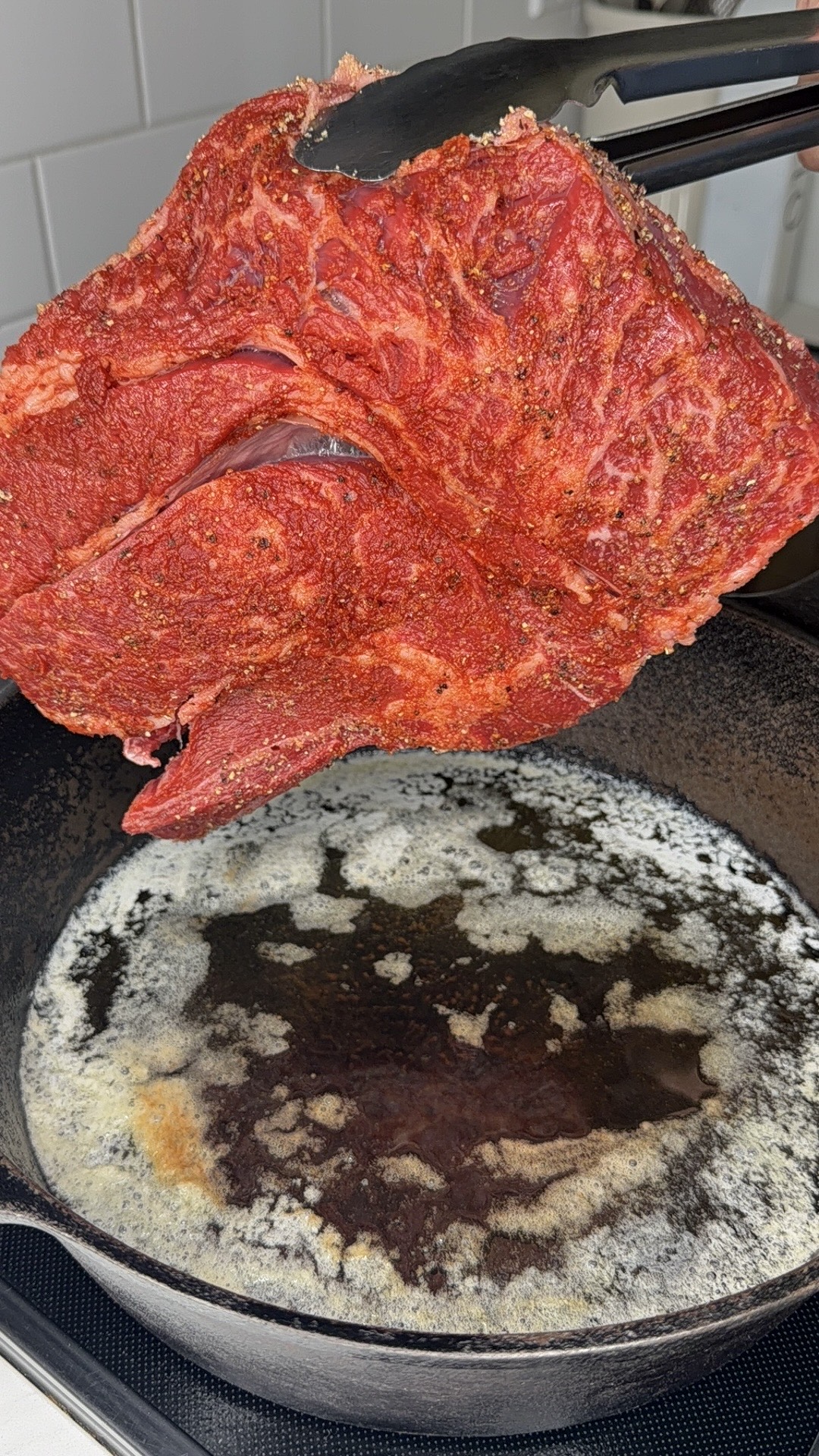 A hand holding metal tongs lowers a large, seasoned raw steak into a hot cast iron skillet with melted butter.