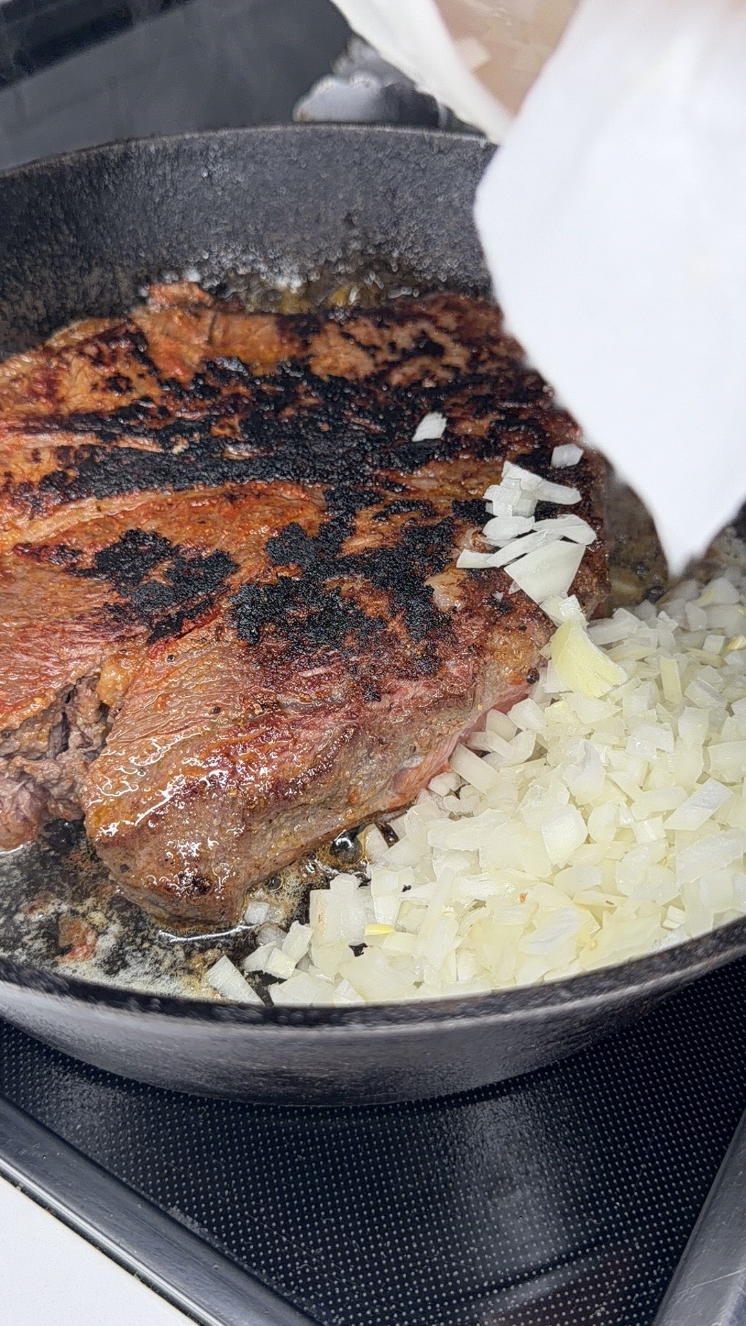 A close-up of a steak searing in a cast iron skillet with diced onions being added beside it on the stovetop.
