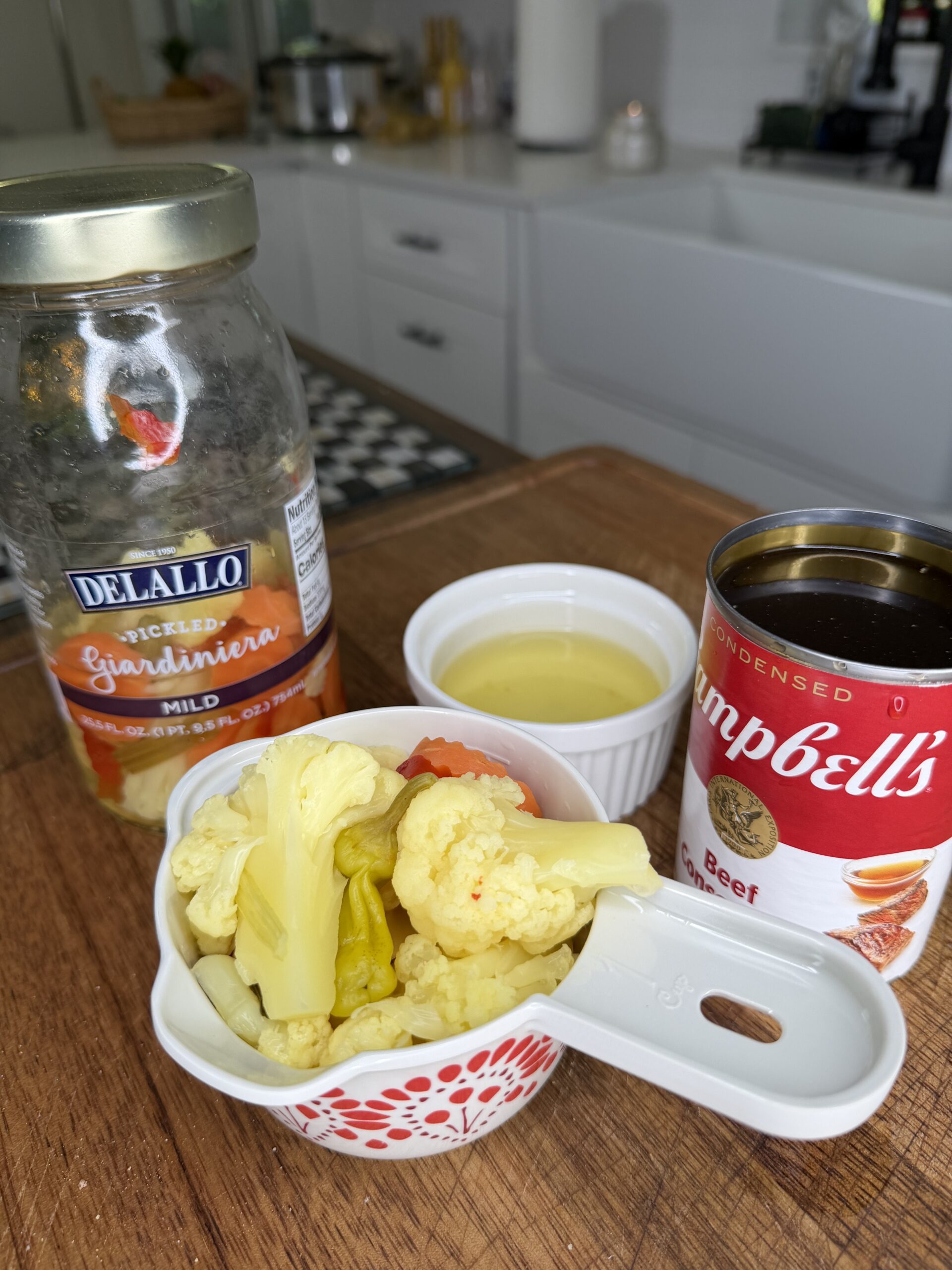 A cutting board with a jar of Delallo pickled giardiniera, a can of Campbell’s beef consommé, a small white ramekin with liquid, and a measuring cup filled with giardiniera vegetables in a kitchen setting.