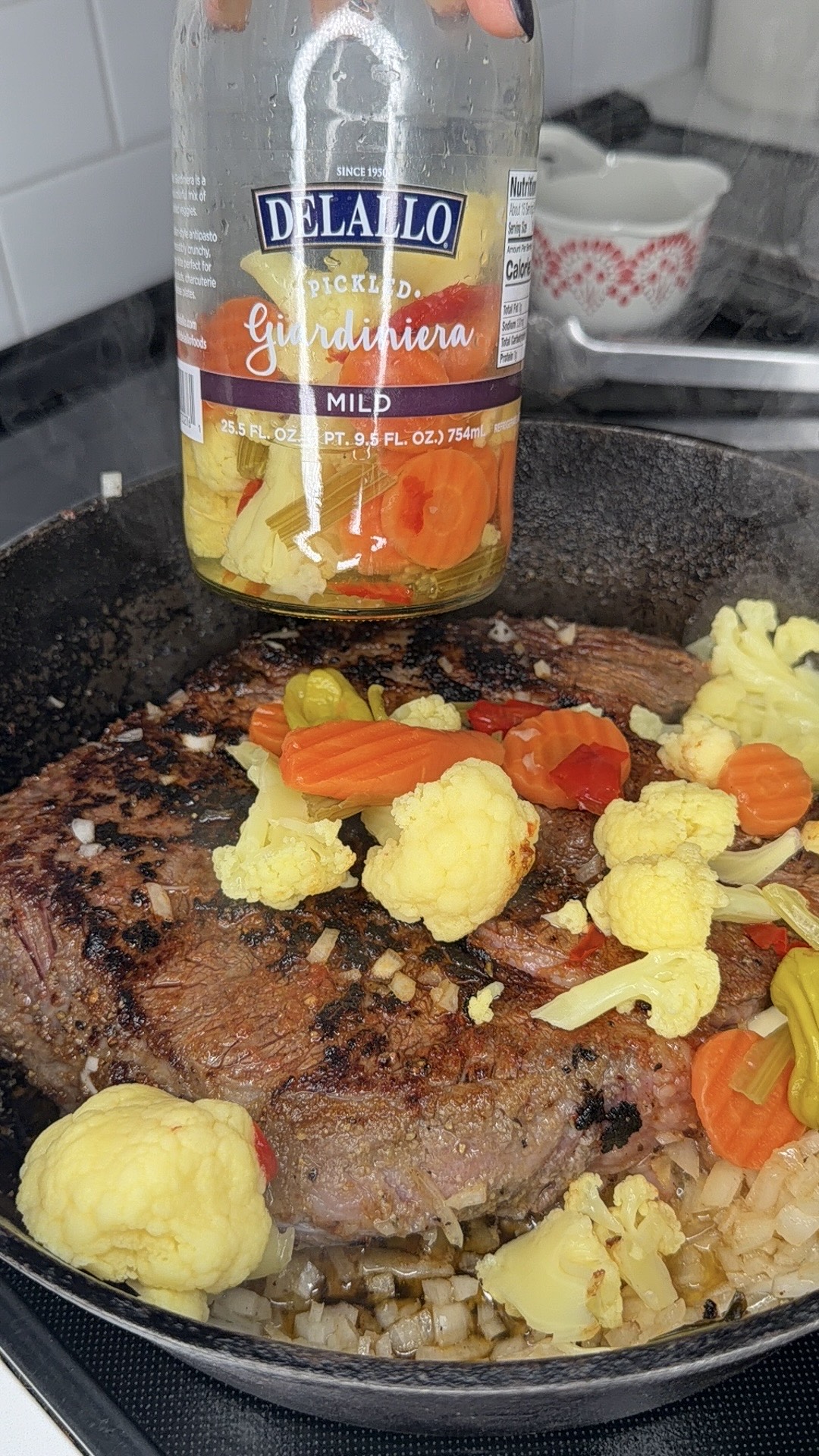 A jar of DeLallo pickled giardiniera is held over a cast iron skillet with a seared beef roast, chopped onions, and pieces of giardiniera vegetables like cauliflower, carrots, and peppers.