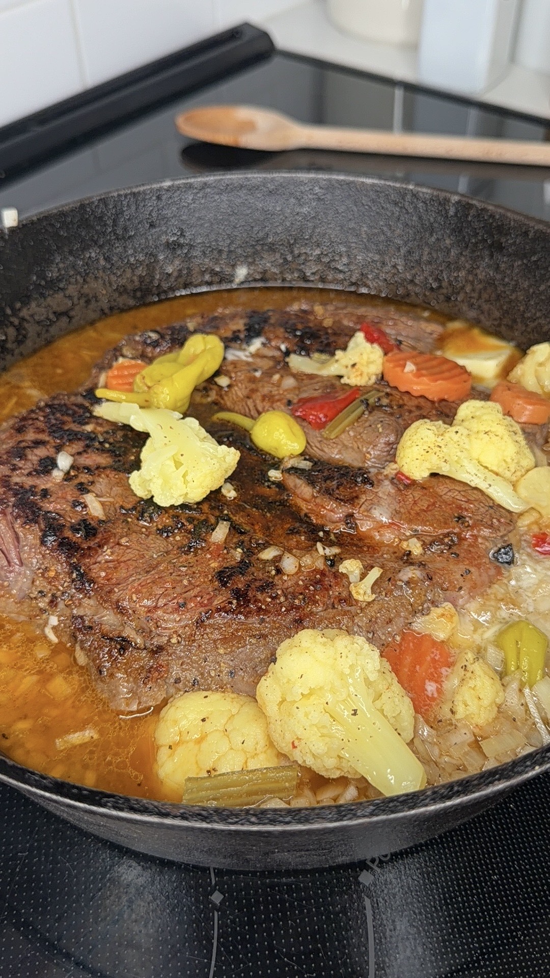 A cast iron skillet on a stove contains a cooked, browned beef roast simmering in broth with cauliflower, carrots, peppers, and seasonings. A wooden spoon rests in the background.