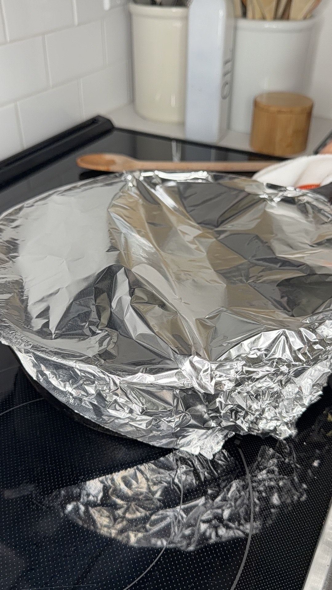 A round baking dish covered with crumpled aluminum foil sits on a black stovetop. Wooden utensils and kitchen containers are visible in the background.
