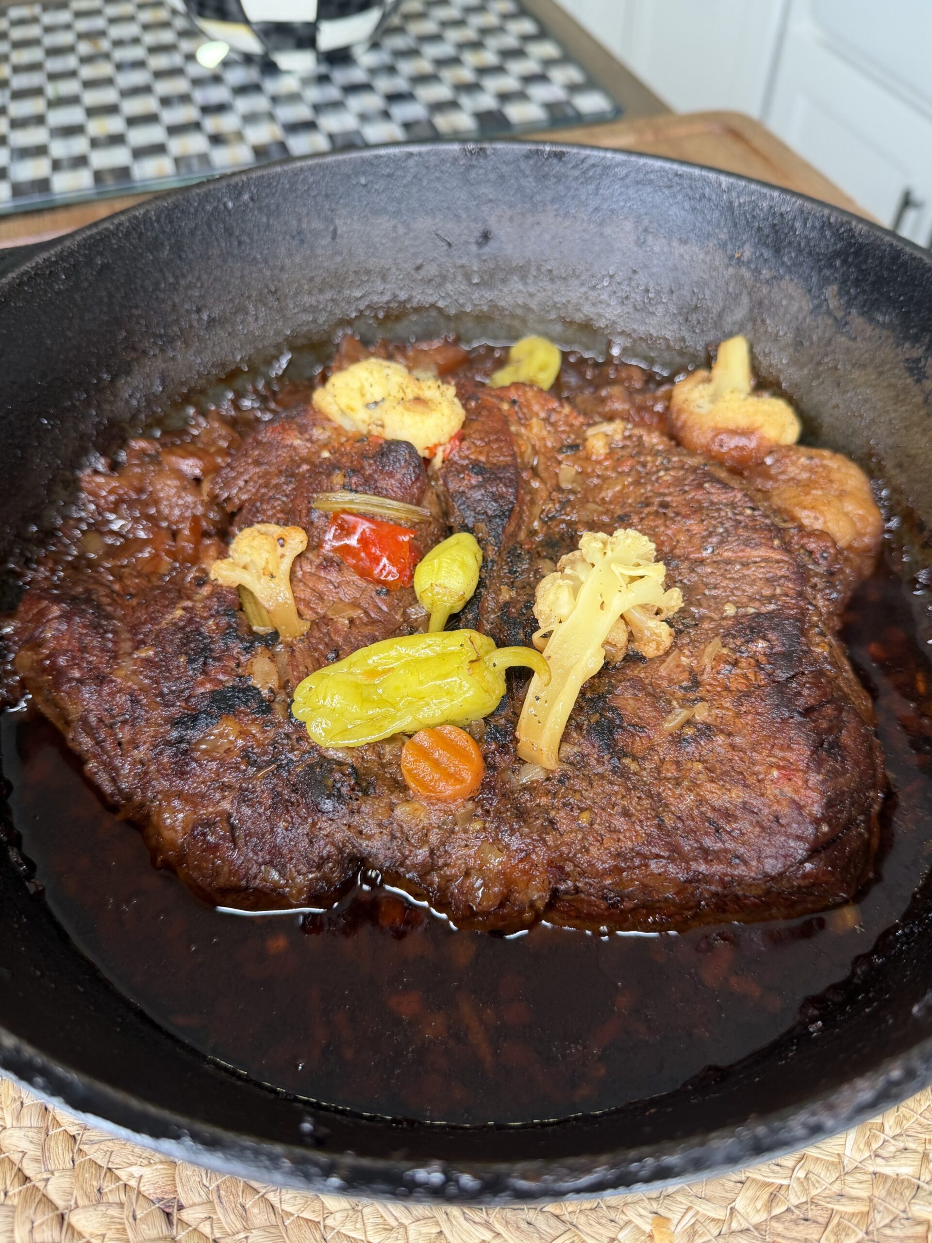 A cooked pot roast sits in a black cast iron skillet, topped with yellow cauliflower florets, peppers, and carrots in a rich, dark sauce. The skillet is on a woven mat, with a checkered dish in the background.