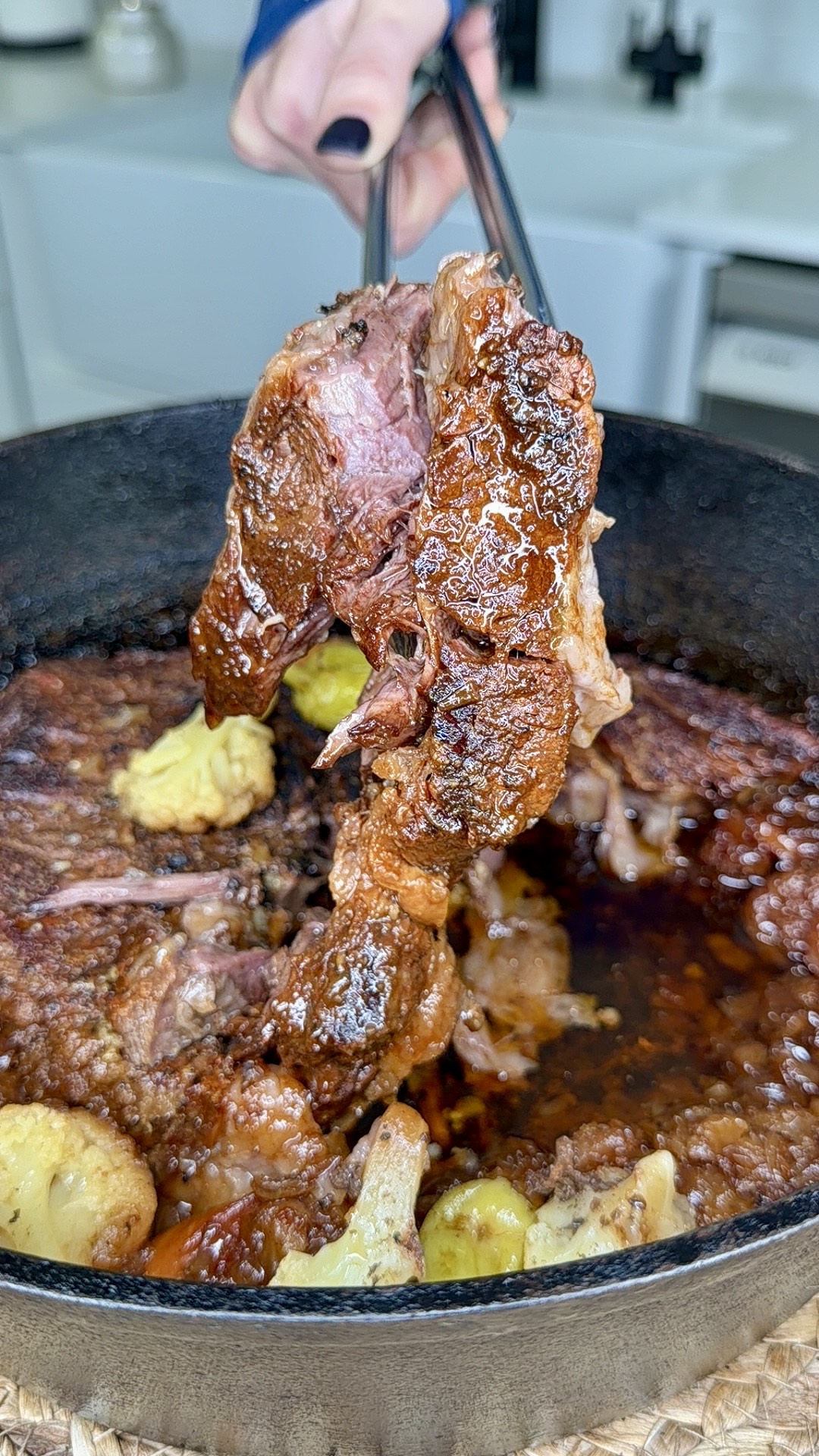A hand lifts a piece of juicy, cooked beef from a cast iron pot filled with tender meat, peppers, and rich juices, with a light kitchen background visible.