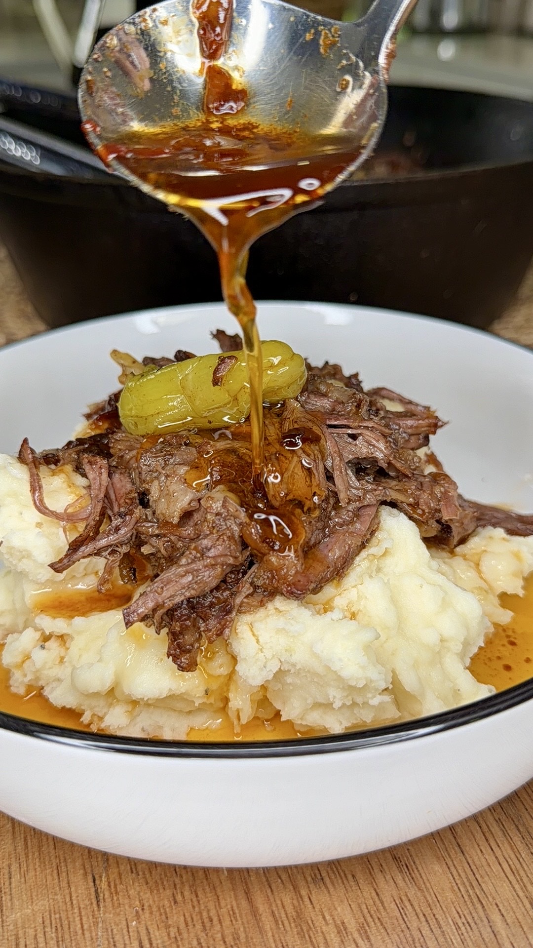 A bowl of mashed potatoes topped with shredded beef and a pepper, with a spoon drizzling rich brown sauce over the dish.