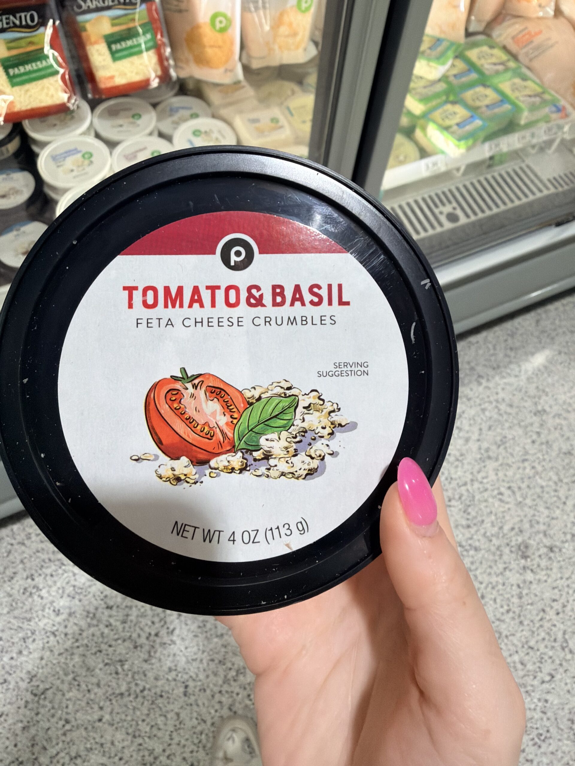 A hand with pink nails holds a container of Publix Tomato & Basil Feta Cheese Crumbles in front of a refrigerated grocery shelf with assorted cheese products.