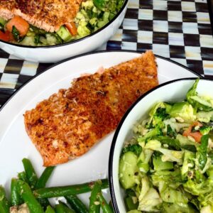 A plate with a seasoned, baked salmon fillet, green beans, and a bowl of chopped salad with lettuce, cucumber, and carrots. In the background, another plate has salmon on top of salad. Everything is on a checkered tabletop.
