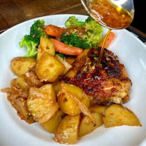 A plate with roasted chicken thigh, golden potatoes, and steamed broccoli and carrots. A spoon drizzles sauce over the chicken. The meal is served on a white plate on a wooden surface.