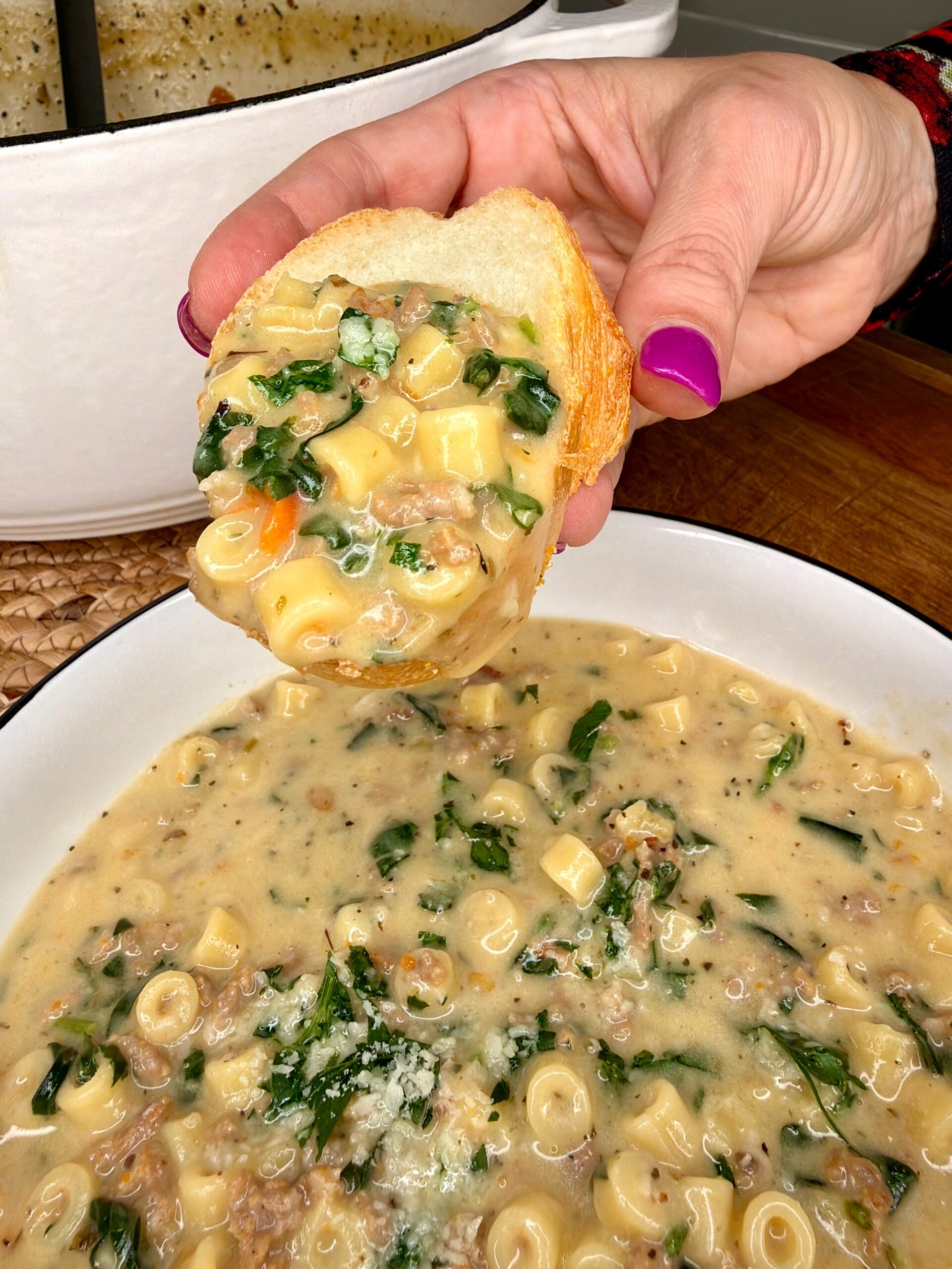 A hand with pink nail polish dips a slice of bread into a creamy soup with pasta, spinach, ground meat, and vegetables, served in a white bowl in front of a large pot.