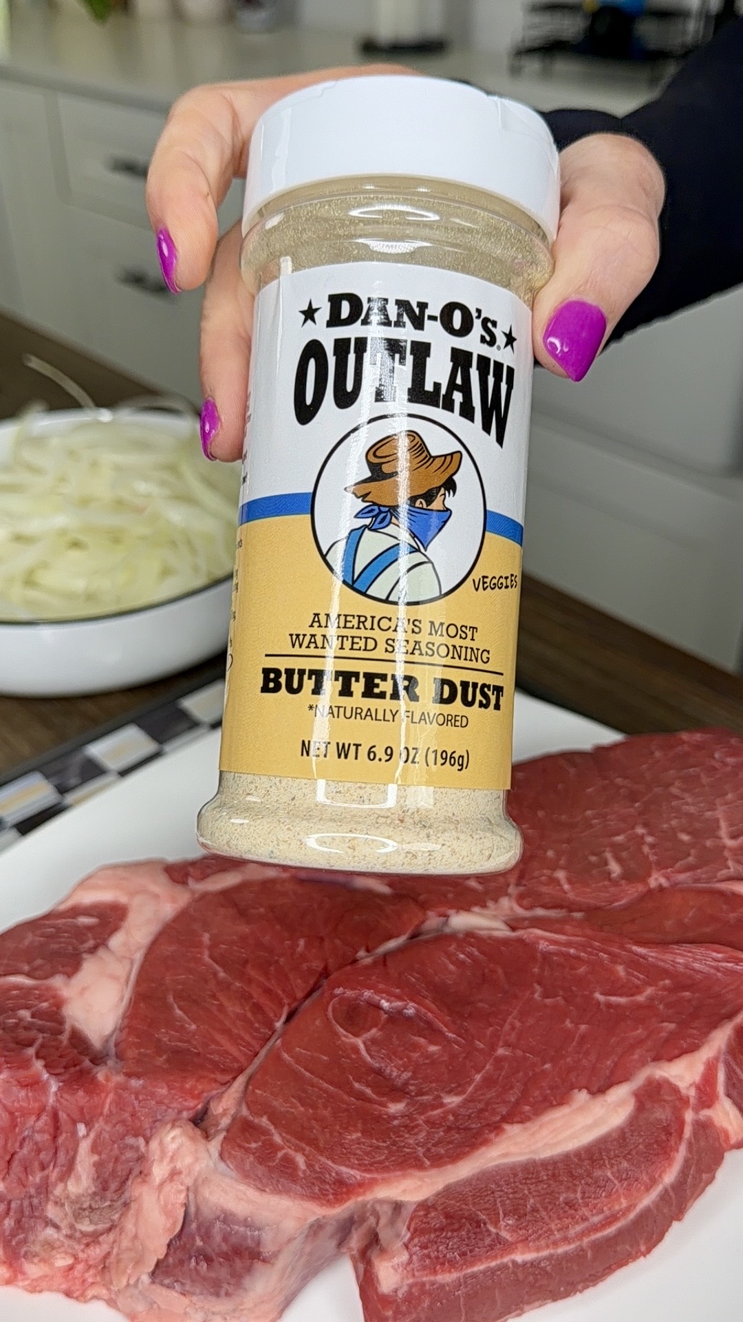 A hand holds a container of Dan-Os Outlaw Butter Dust seasoning above a raw steak on a plate, with sliced onions and a knife visible in the background.