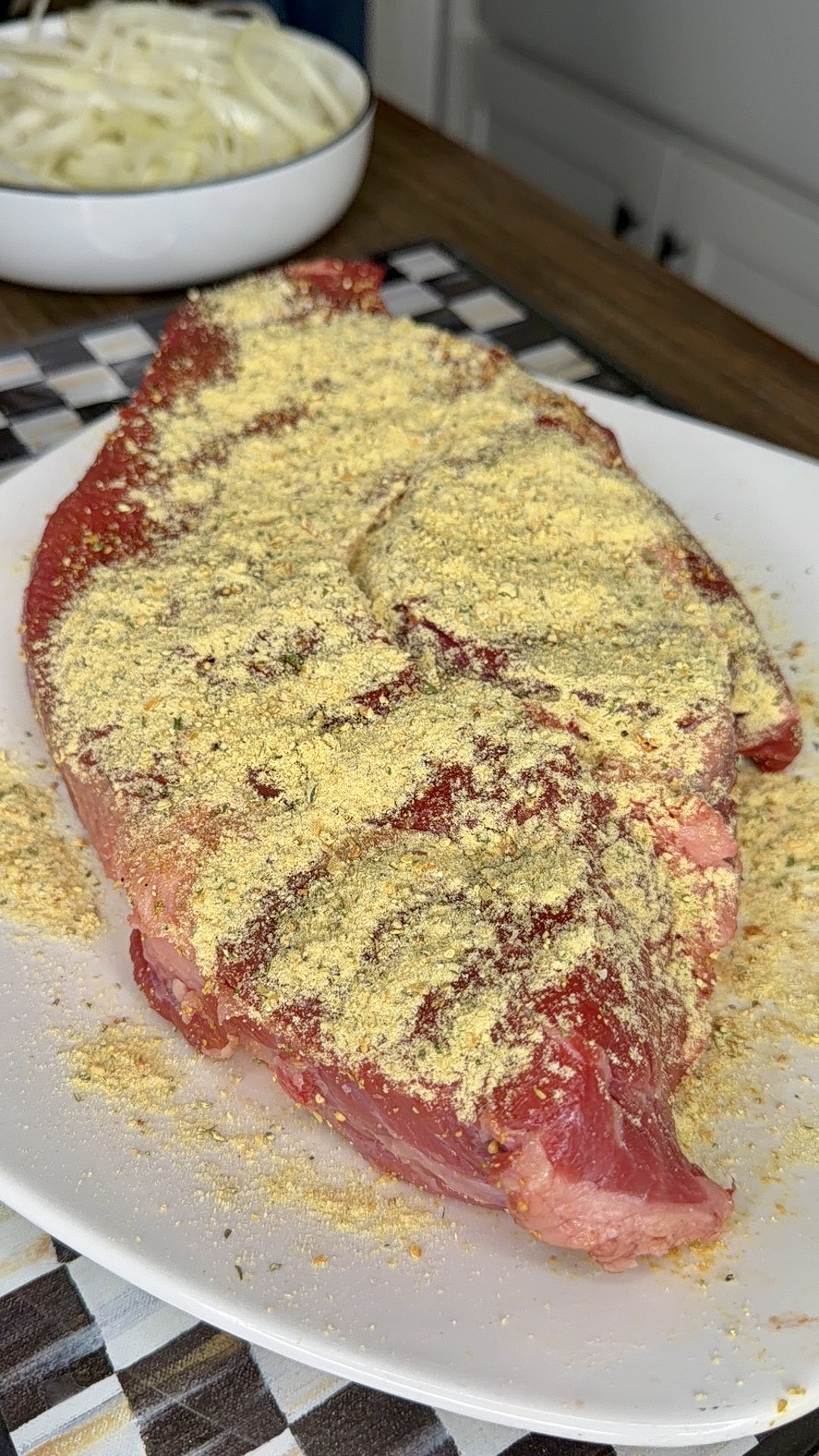 A raw steak covered with a generous layer of yellow seasoning sits on a white plate. In the background, there is a bowl filled with sliced onions on a checkered placemat.