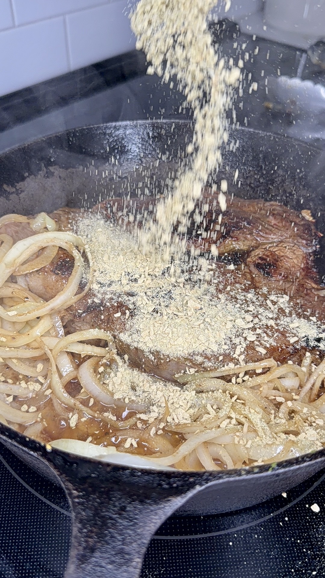 Sliced onions and steak cooking in a cast iron skillet as a powdered seasoning is being sprinkled over them, creating a savory, flavorful dish.