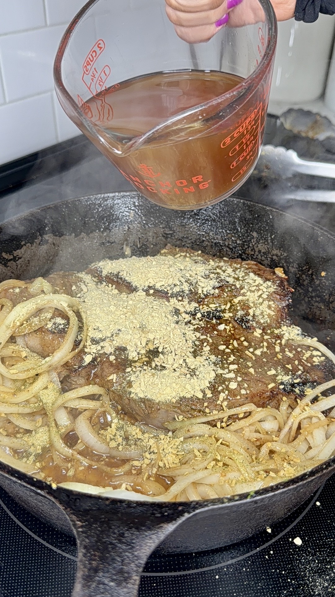 A hand pours broth from a glass measuring cup into a cast iron skillet filled with sliced onions, seasoning, and browned meat, all sizzling on a stovetop.