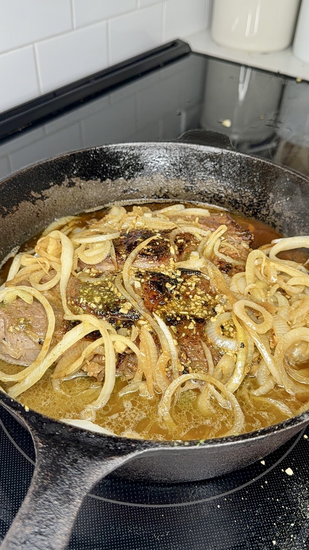 A cast iron skillet on a stovetop filled with cooked meat, sliced onions, and broth. The meat is browned and topped with seasonings, while the onions appear softened and lightly caramelized.