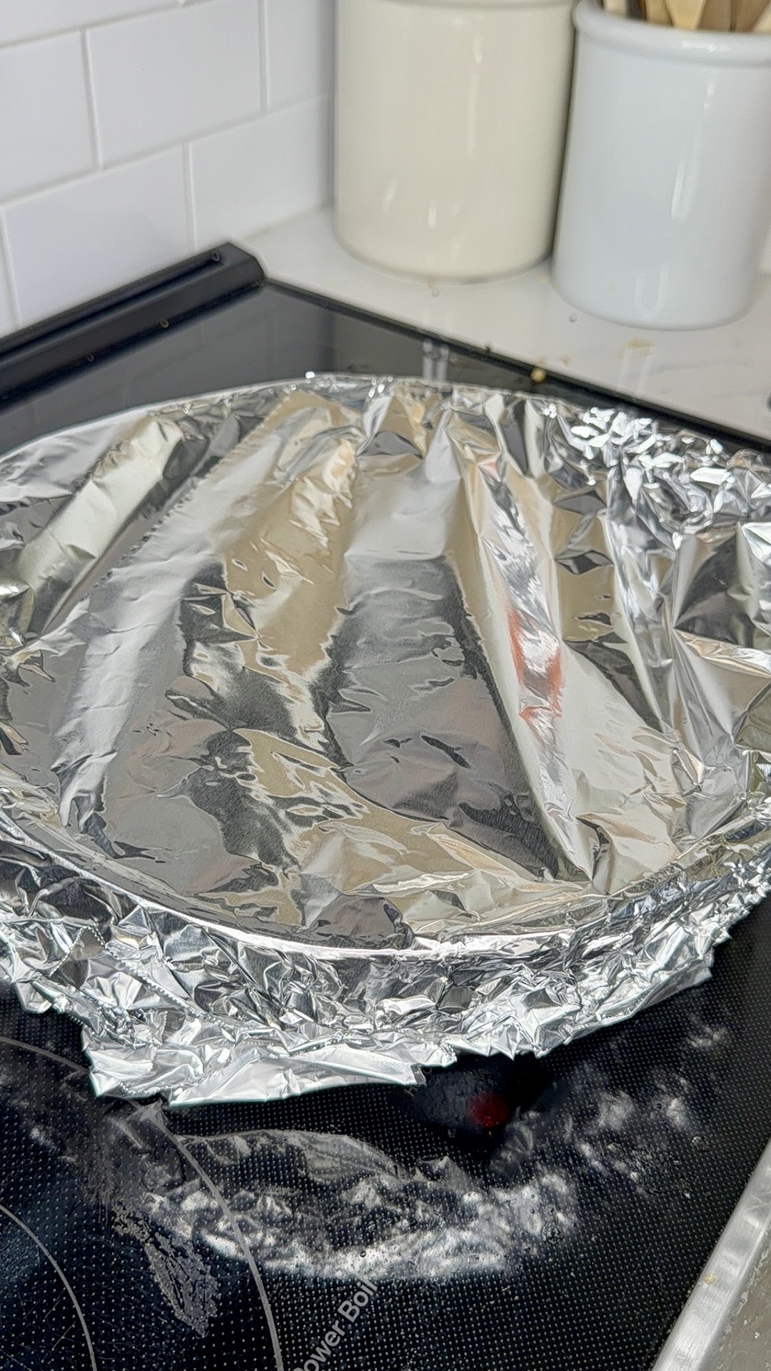 A round baking dish covered with crinkled aluminum foil sits on a stovetop, with some foil overlapping the edges. Kitchen canisters and utensils are visible in the background.