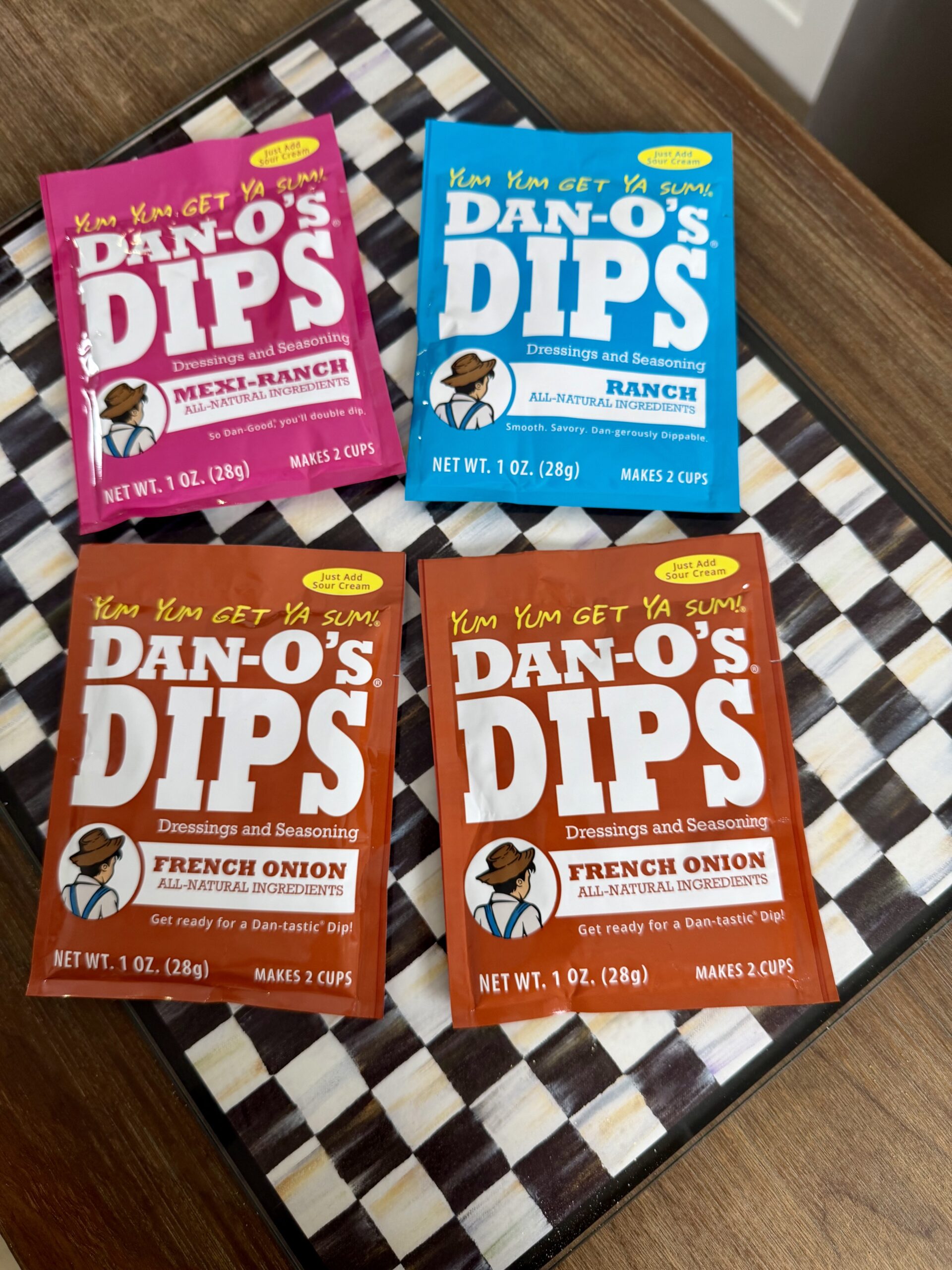 Four packets of Dan-O’s Dips seasoning are arranged on a checkered surface. Flavors include two French Onion (orange), one Cool Ranch (blue), and one Spicy Meat-Ranch (red). Each packet makes two cups.