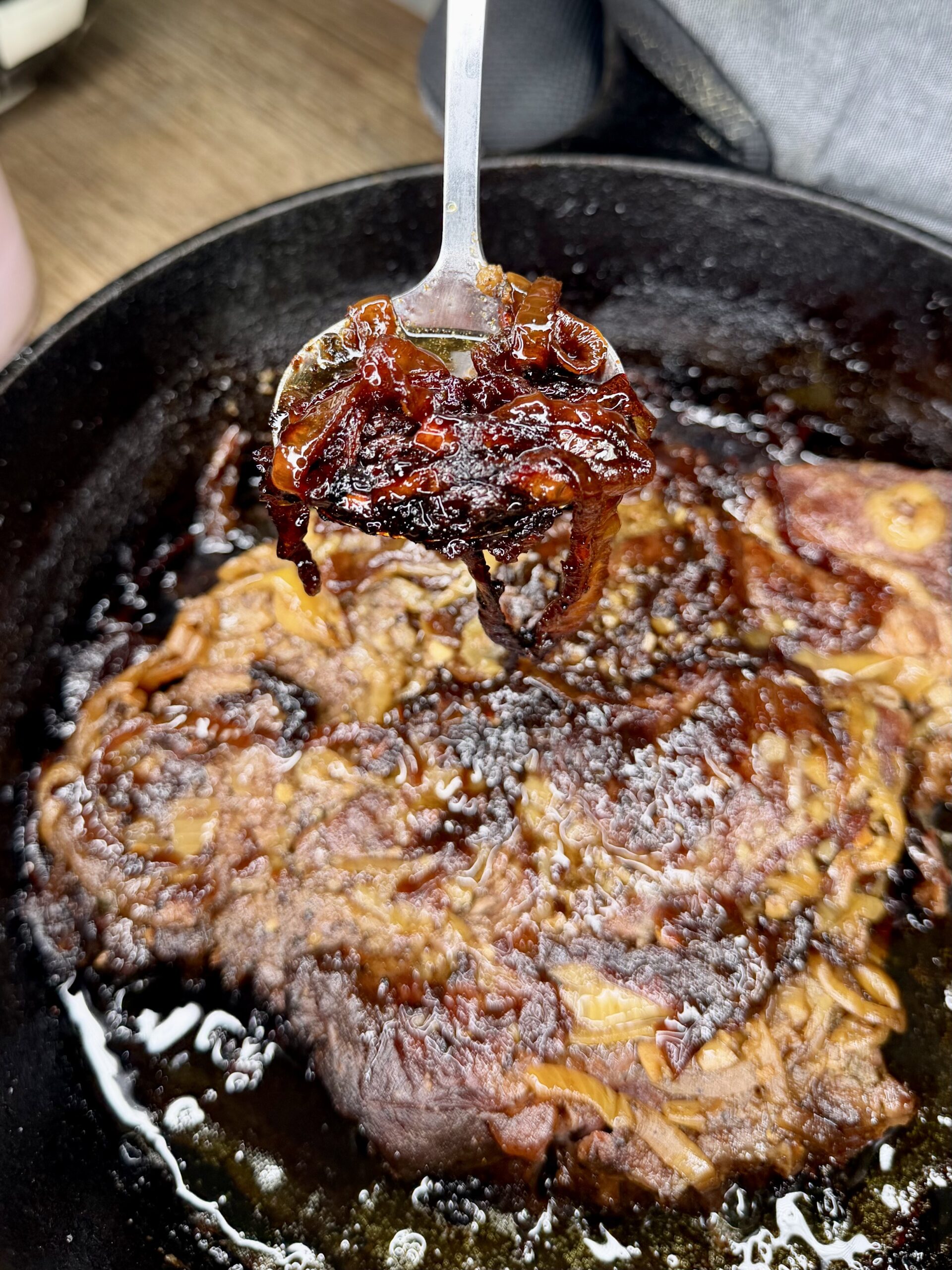 A close-up of caramelized onions being lifted with a spoon over a cast iron skillet filled with browned, sizzling meat and juices.