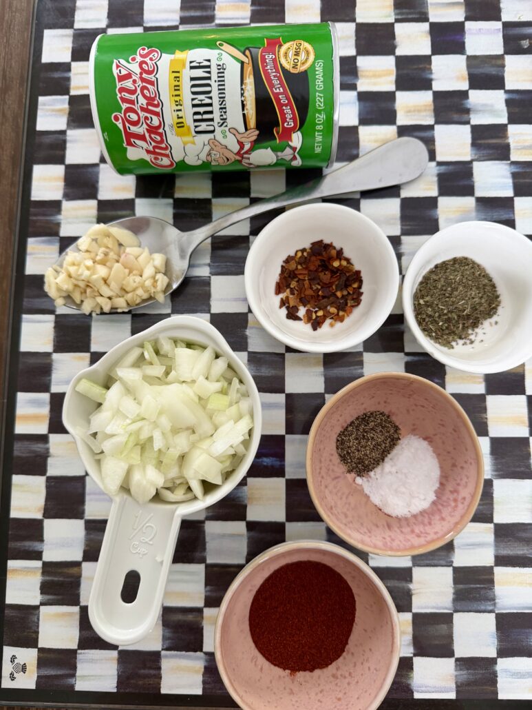 A can of Tony Chachere’s Creole Seasoning, chopped garlic, crushed red pepper, dried oregano, chopped onions, black pepper, salt, and paprika are arranged in small bowls on a checkered surface.